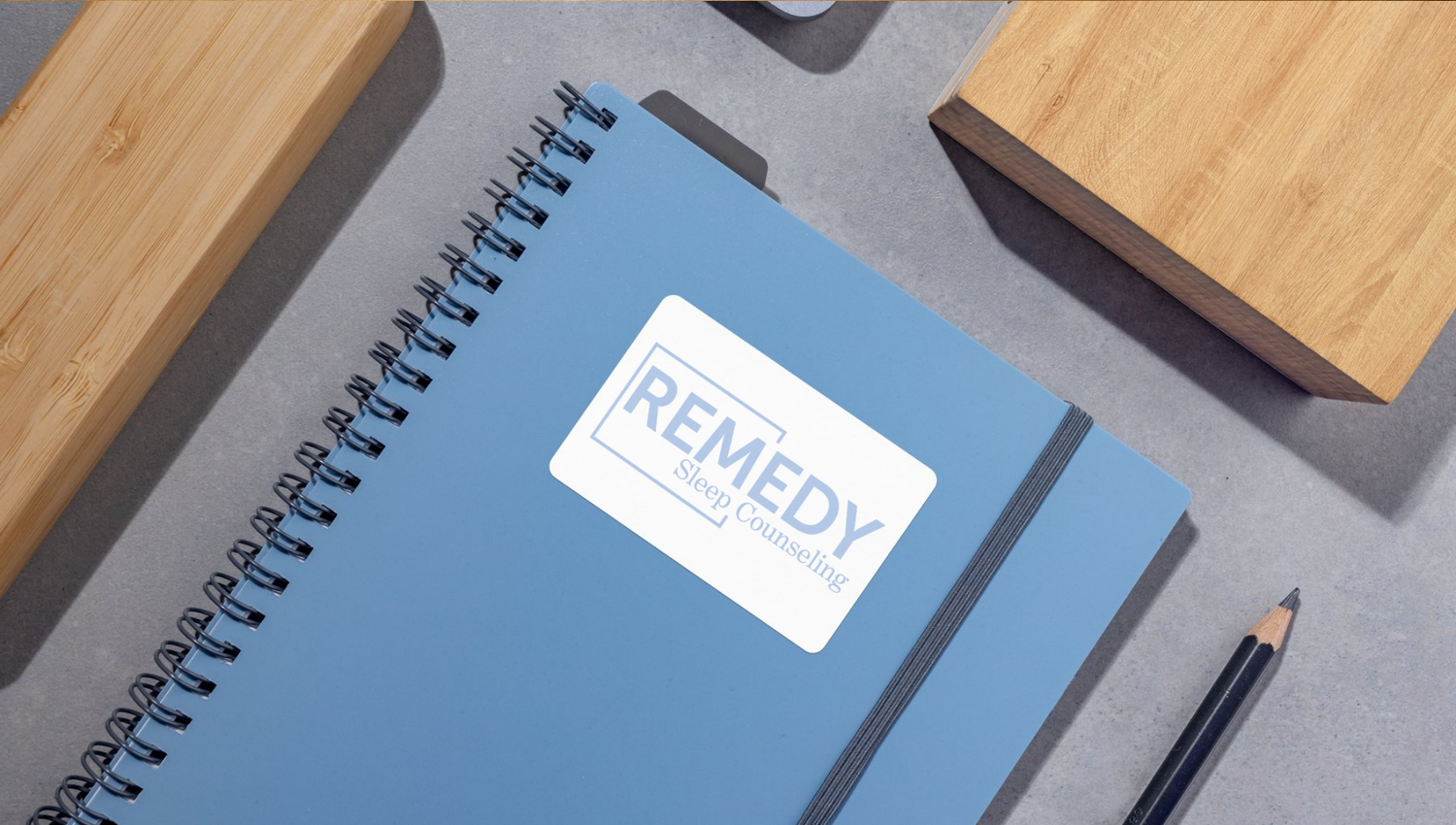 Spiral notebook with blue cover and white text lies on wooden surface next to black pen and other office supplies.
