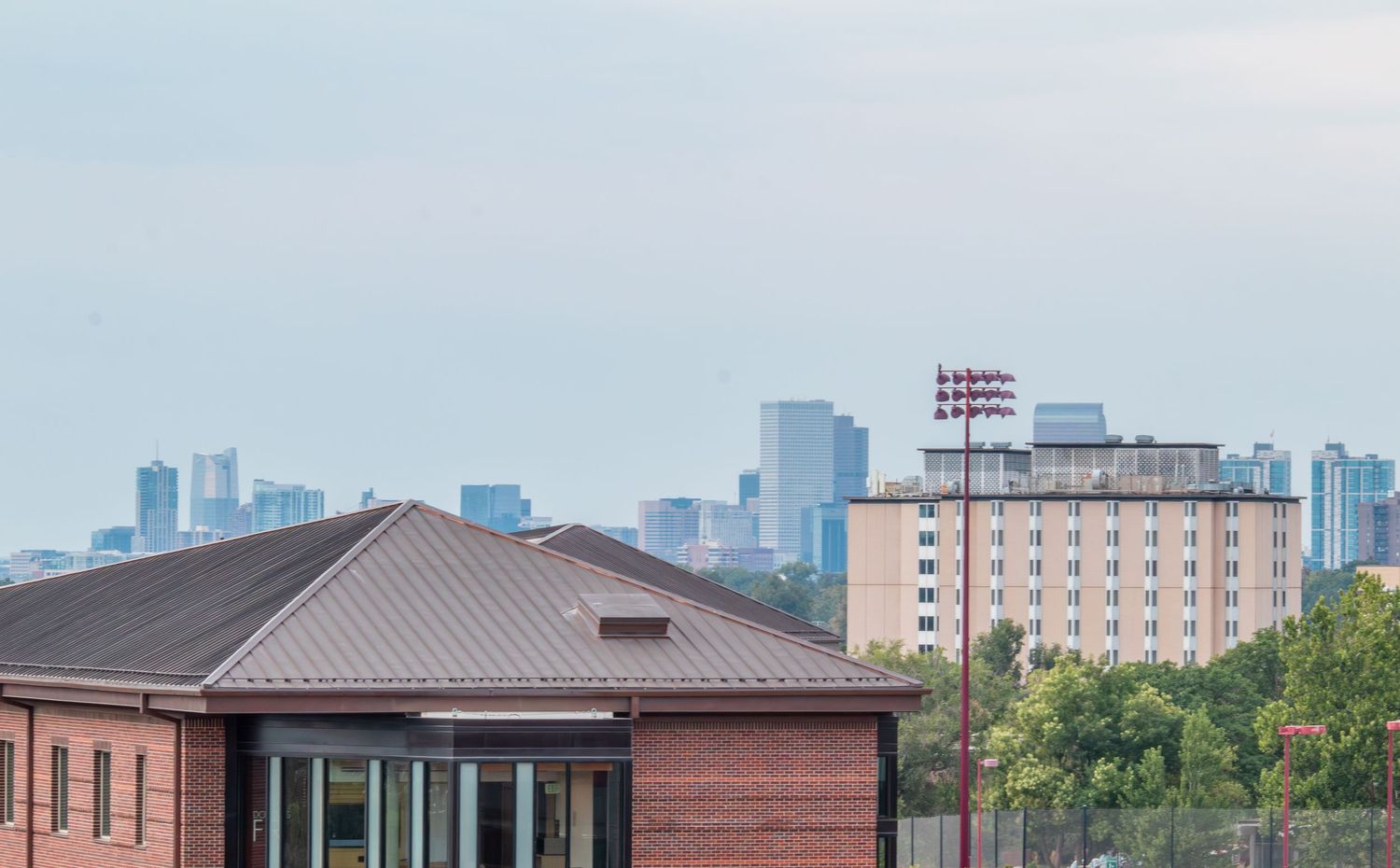 Urban skyline view behind a brick building with metal roof and red light tower on a hazy day.