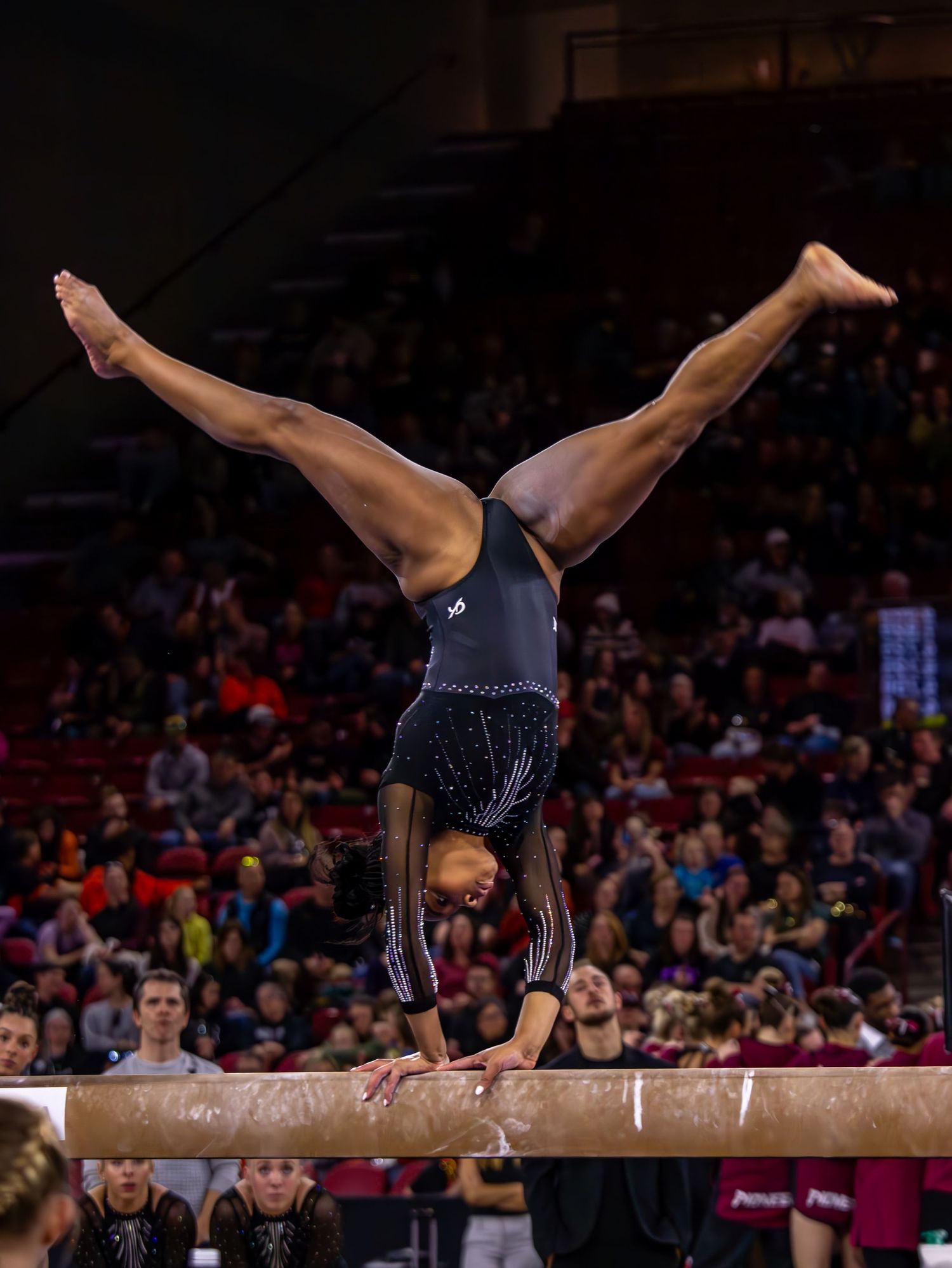 Gymnast performing a handstand split on the balance beam during a competitive gymnastics meet.