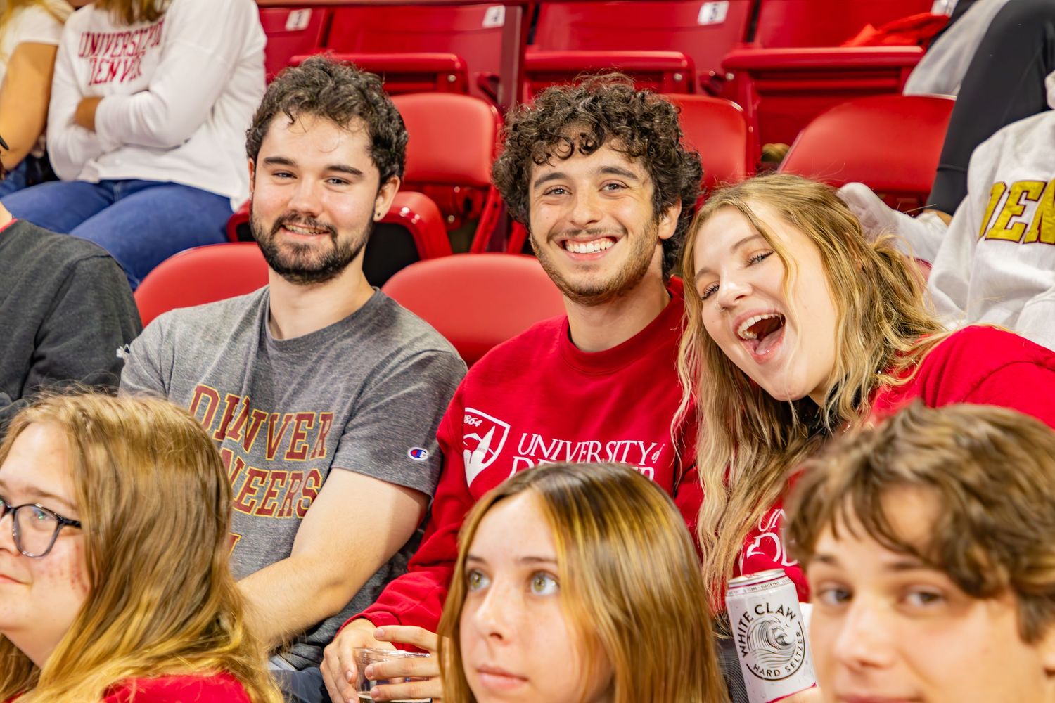Three smiling college students in red university shirts enjoying a sports game in stadium bleachers.