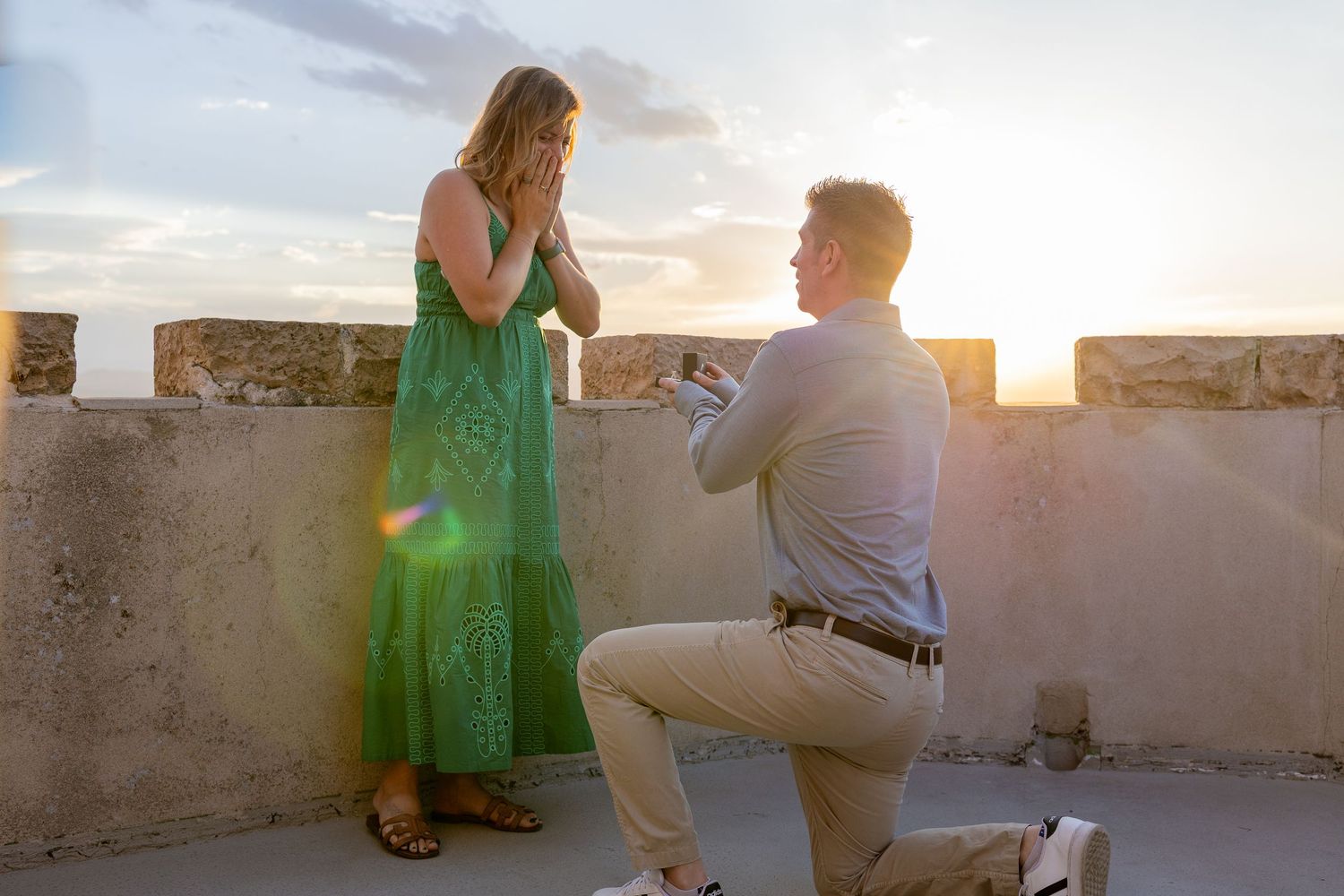 Man proposes on rooftop at sunset while woman covers her face with hands in emotional surprise.