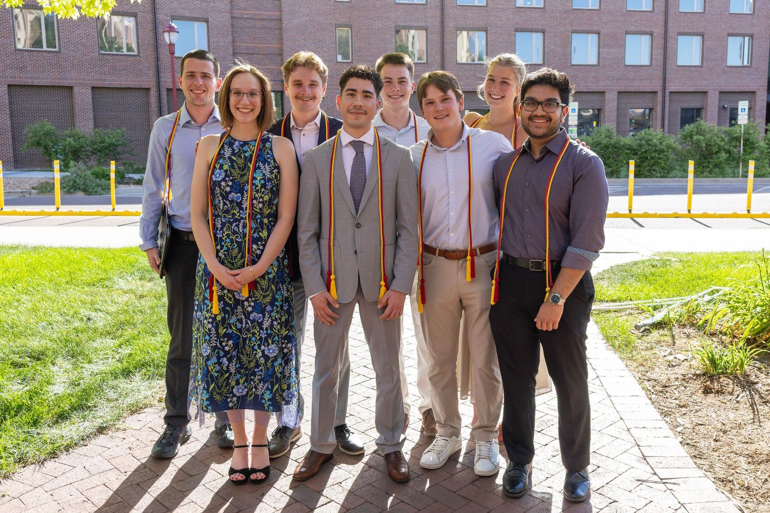 Group of eight college graduates smiling outdoors, wearing graduation stoles, in front of a brick building.