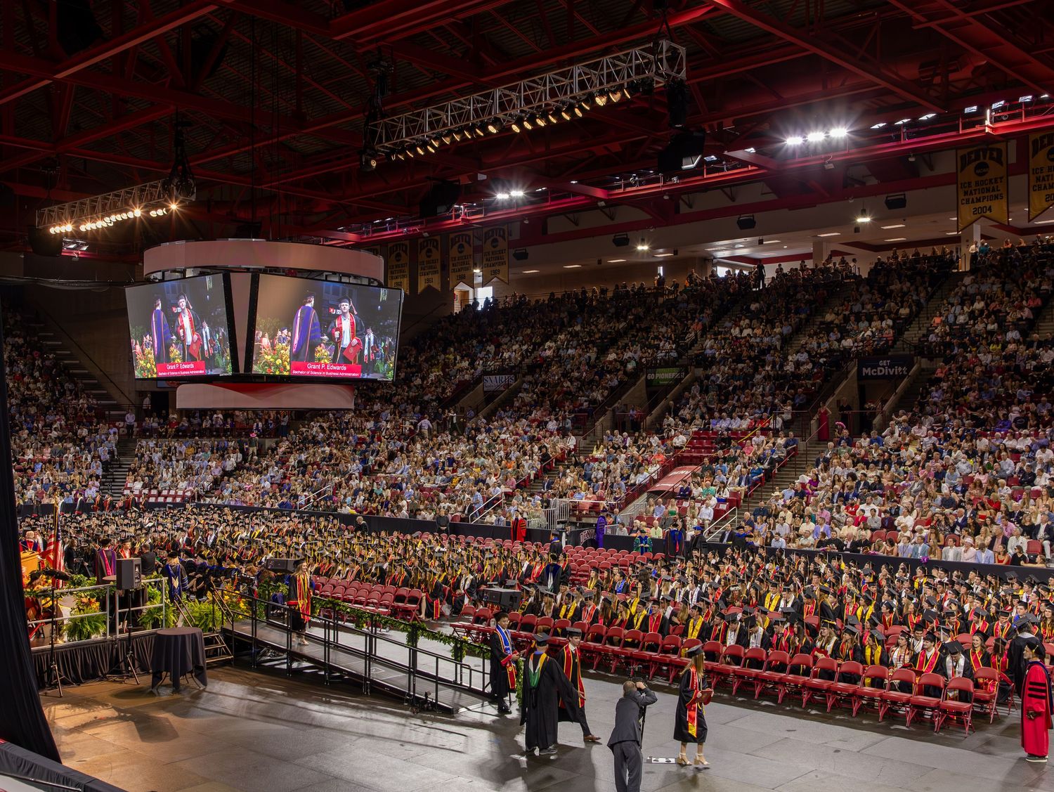 Large indoor graduation ceremony with packed arena, red-robed graduates and big video screens displaying the event.