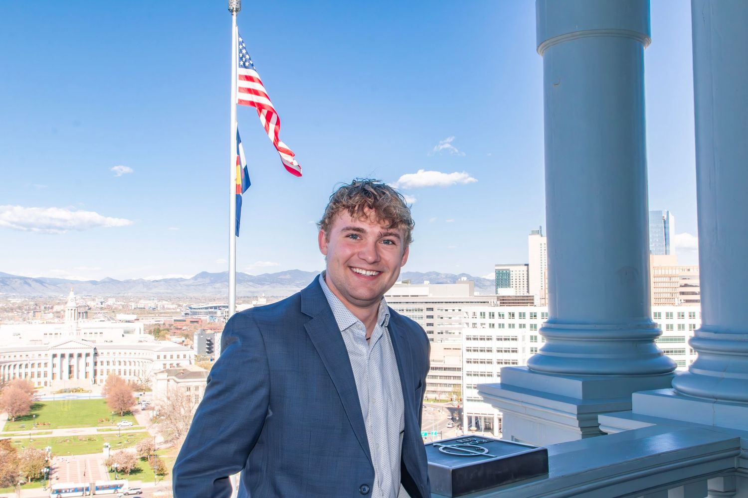 Young man in blue blazer smiling on a balcony with US flag, Colorado Capitol building and mountains in background.