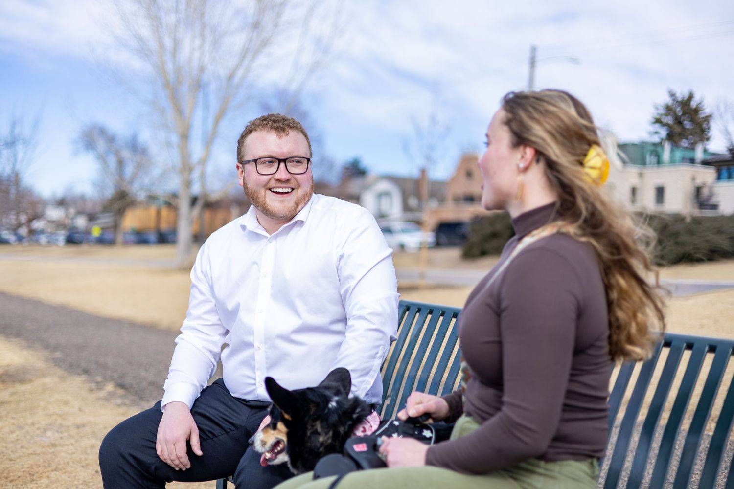 A couple sits together on a park bench during an outdoor photo session on a sunny day.