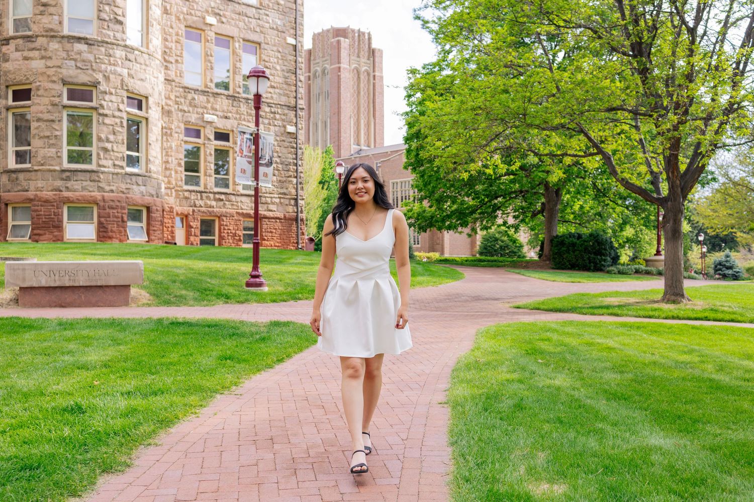 A young woman in a white dress walks along a brick path on a lush college campus with stone buildings.