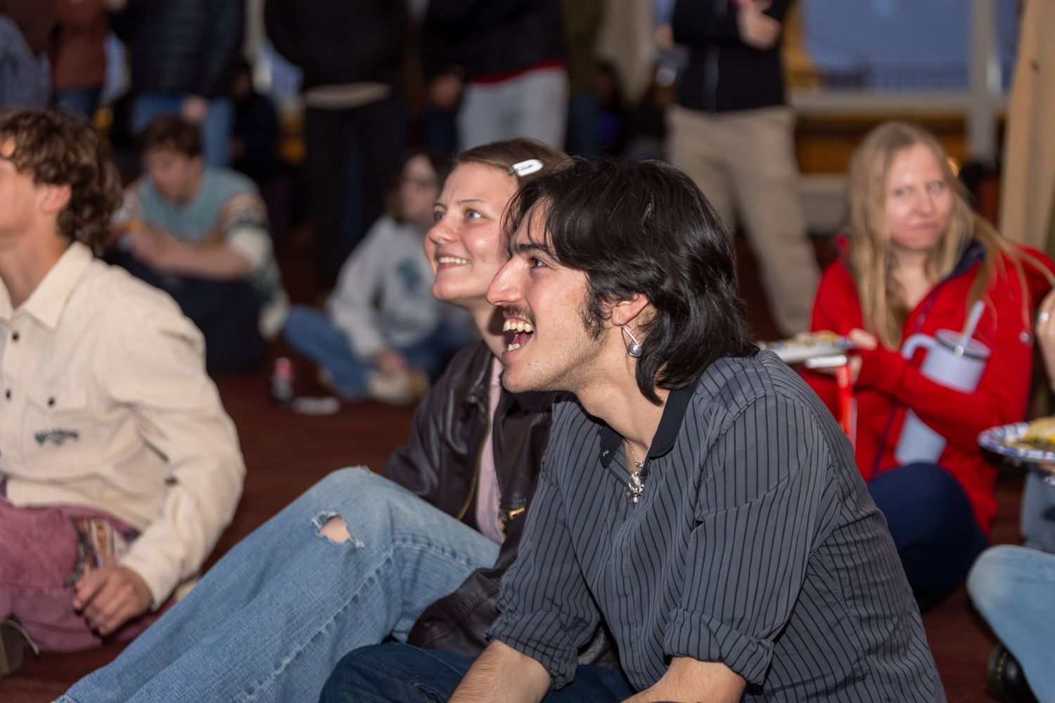 Group of young adults sitting on floor laughing and enjoying an indoor event together.