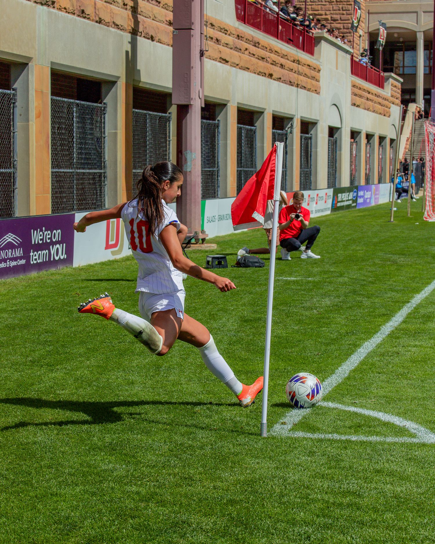 Female soccer player in white uniform takes a corner kick on a green field during a college match.