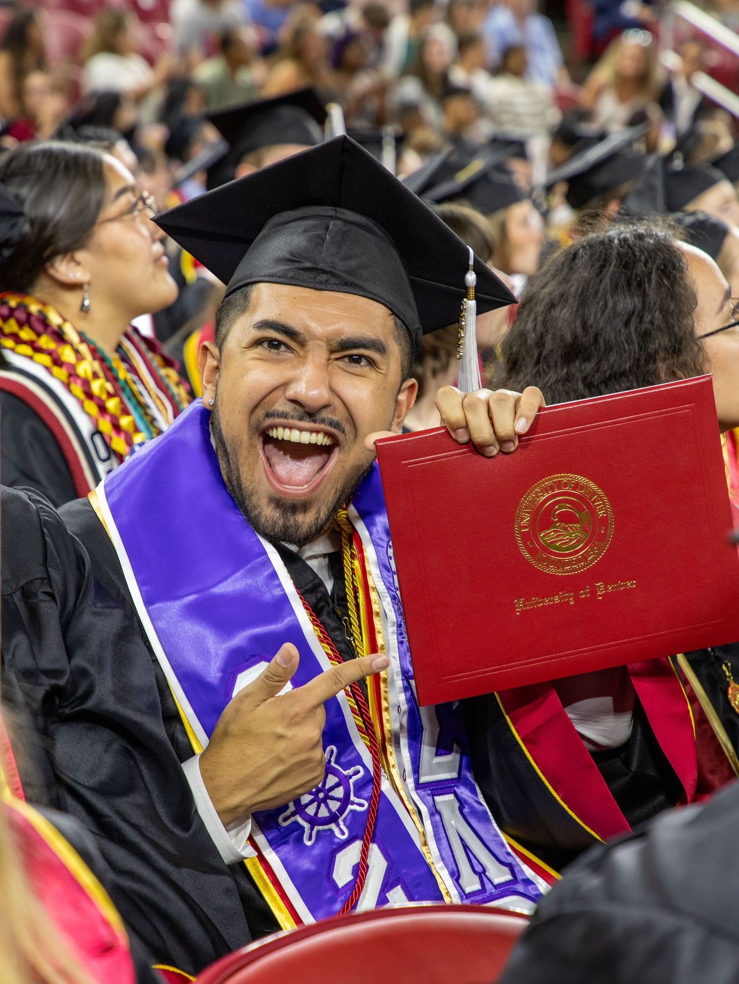 Excited male graduate holds red diploma cover while wearing cap, gown, and purple sash at graduation.
