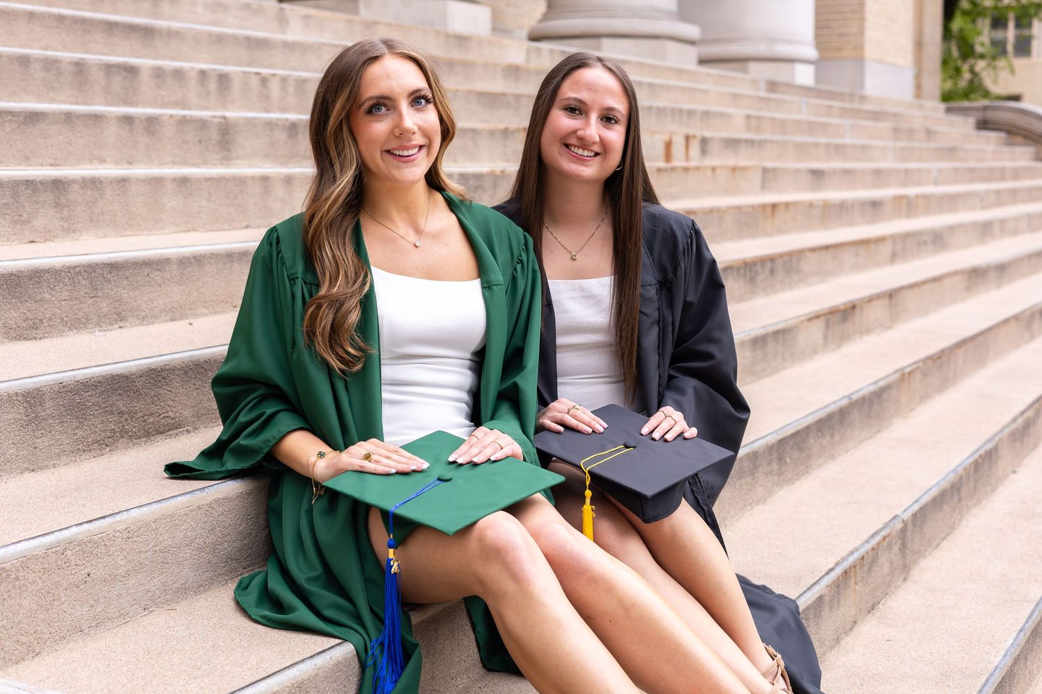 Two smiling college graduates in green and black graduation gowns sitting on stone steps holding caps.