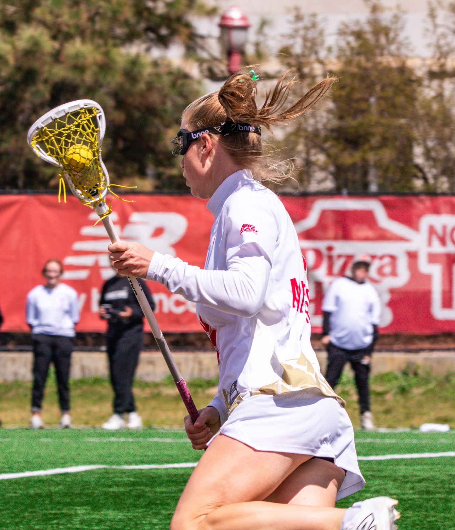 Female lacrosse player in white uniform running with stick during outdoor game on turf field.
