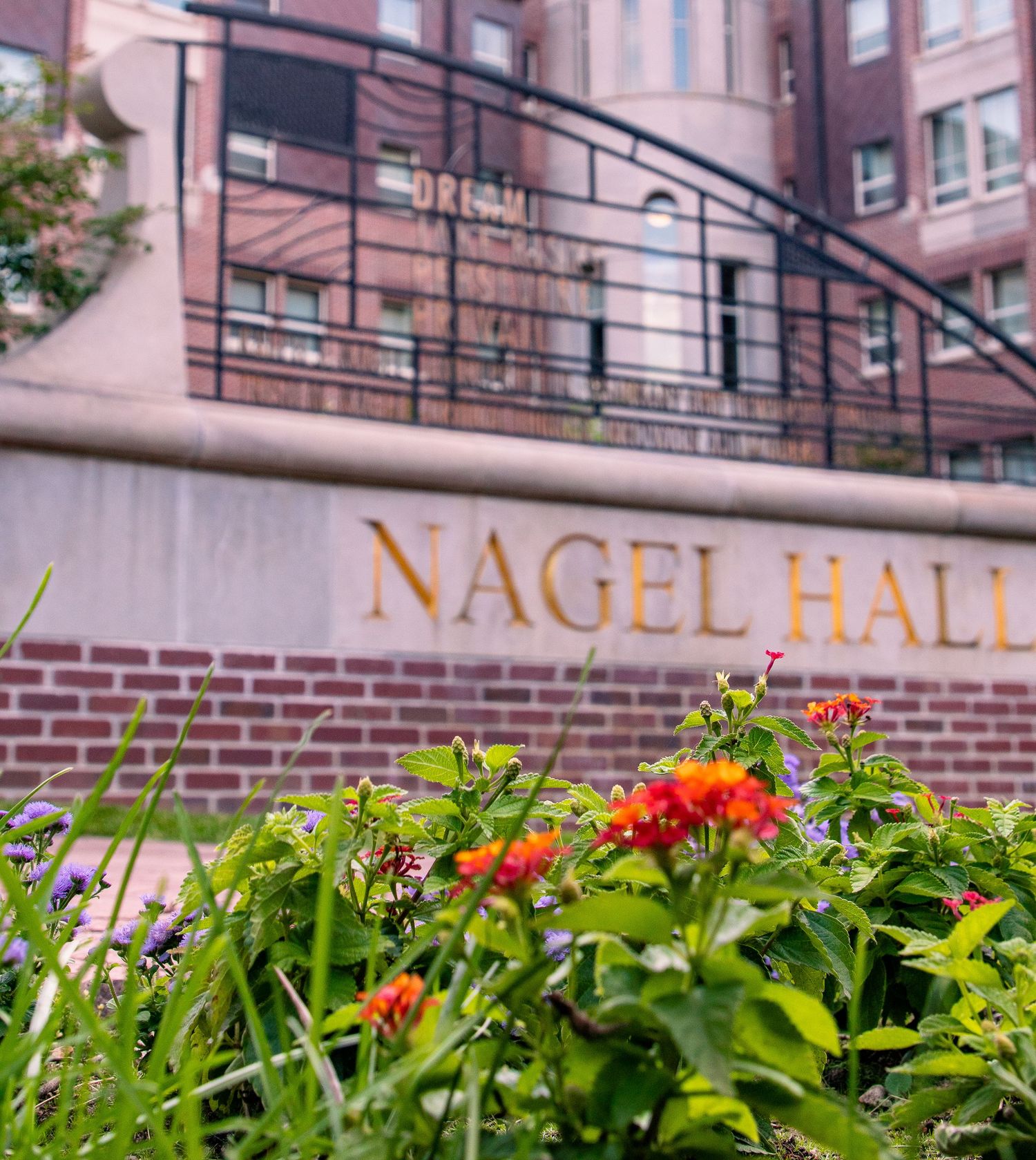 Nagel Hall brick entrance sign with colorful lantana flowers blooming in the foreground on a college campus.