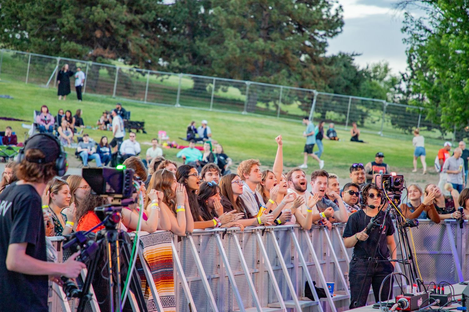 Excited concert crowd presses against barrier at outdoor University of Denver campus music event at dusk.