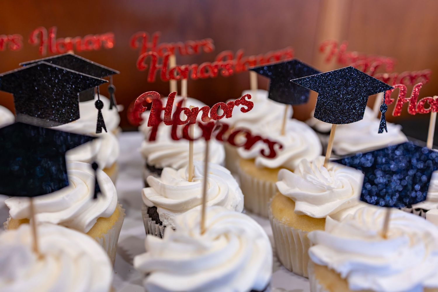 White frosted cupcakes decorated with graduation cap toppers and red glittery Honors signs for a graduation celebration.