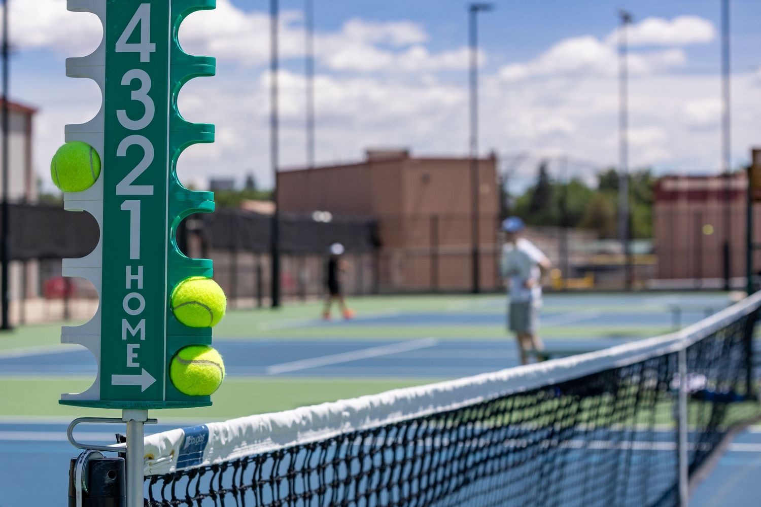 Tennis court with score tracker holding yellow balls near the net, players visible on blue courts in background.