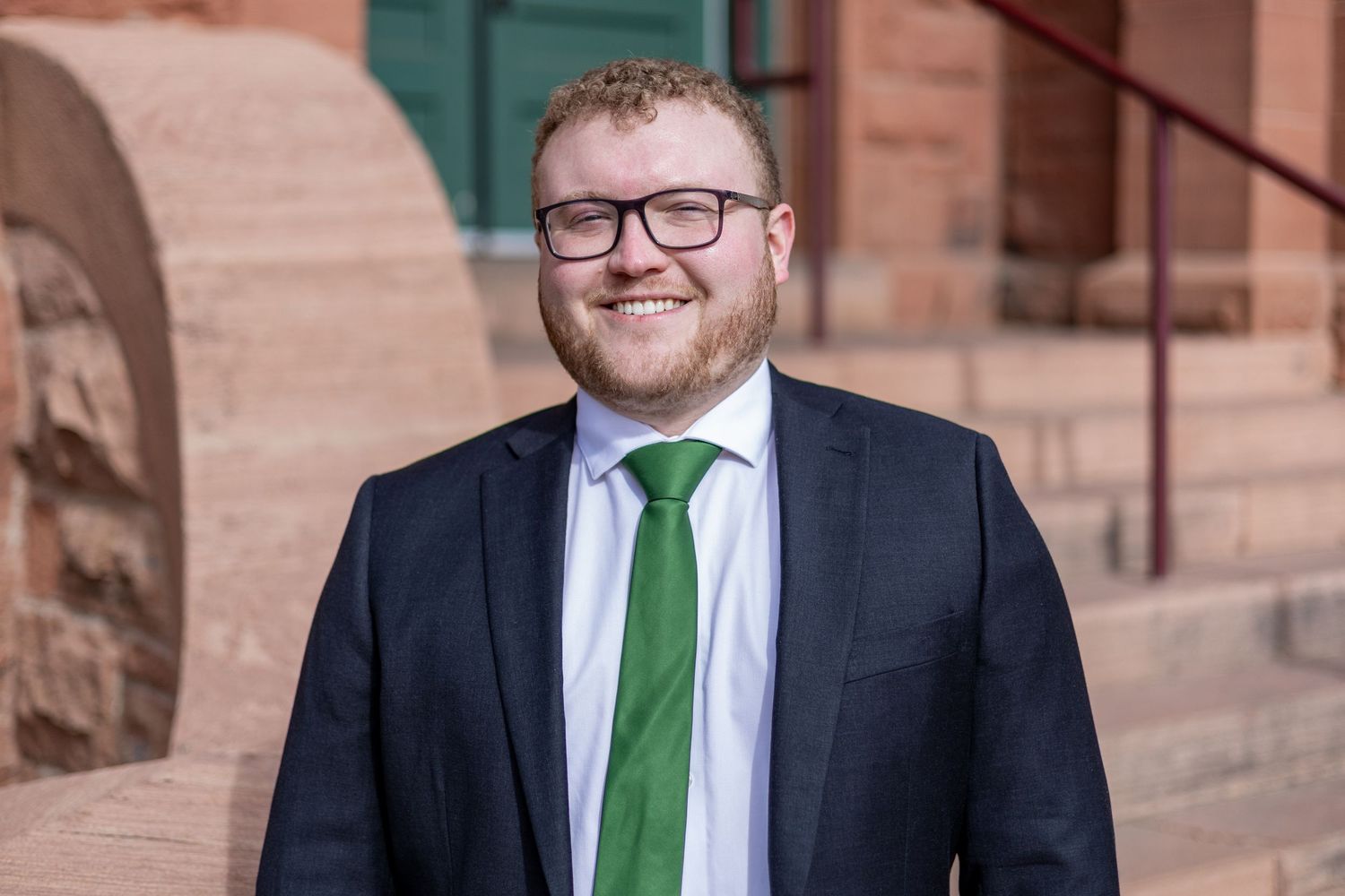 Professional wearing a black suit with green tie and glasses poses against a brick wall in a series of photos.