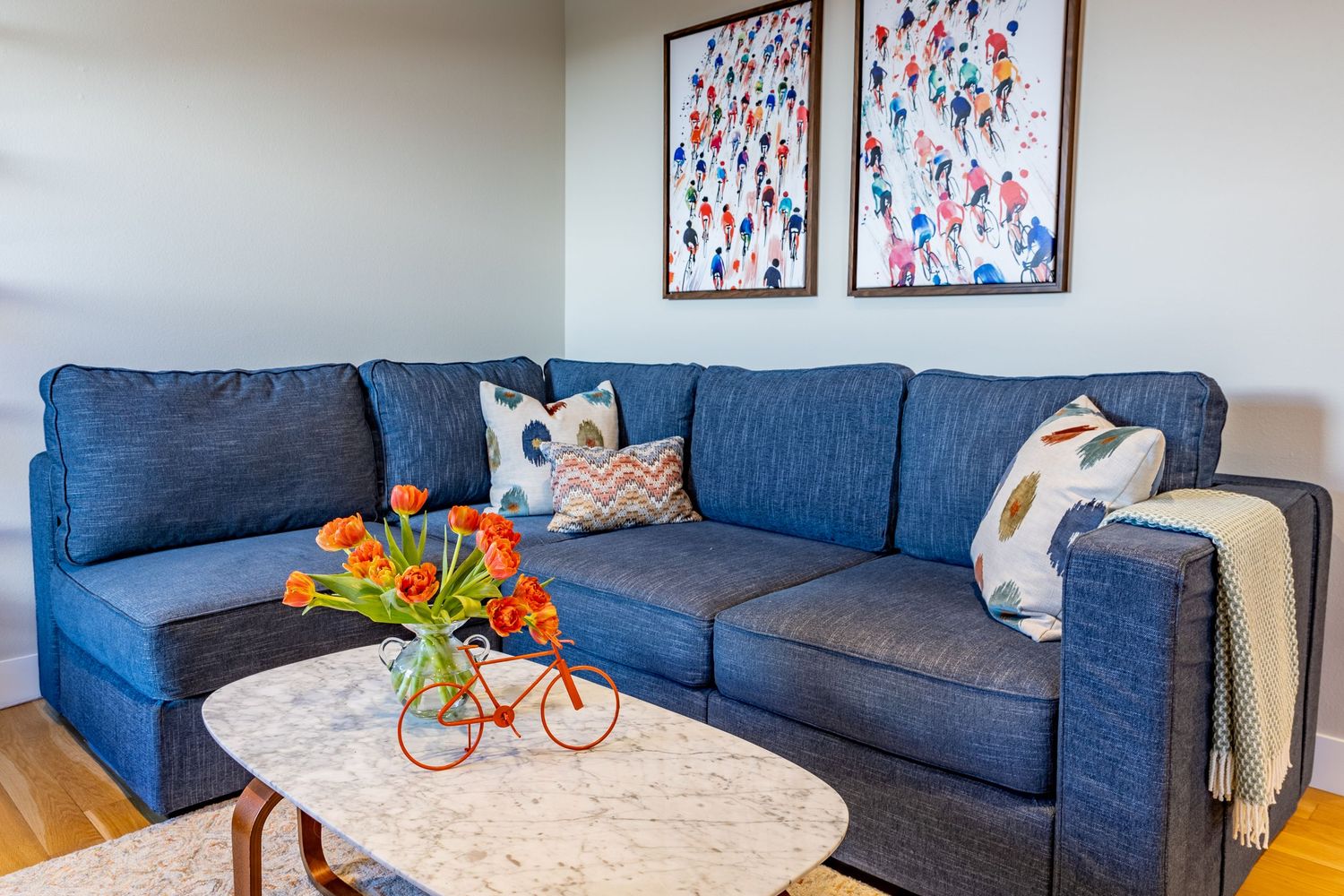 A modern living room features a blue sectional sofa, round marble coffee table, and colorful wall art above.