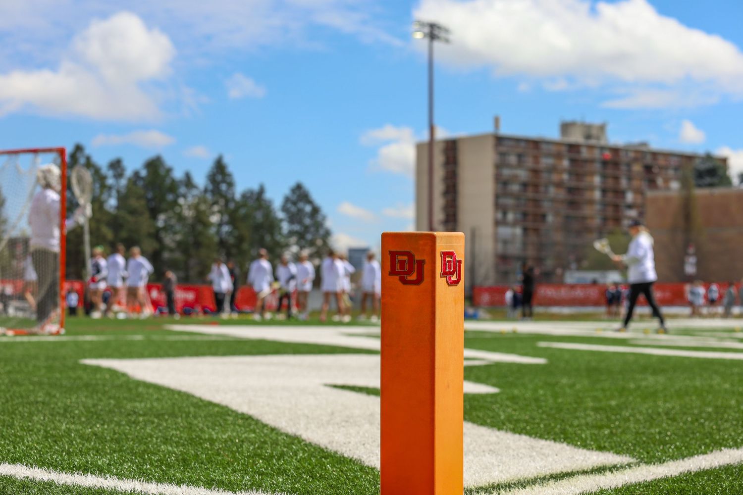 Orange goal line pylon on artificial turf football field with players and buildings visible in background.
