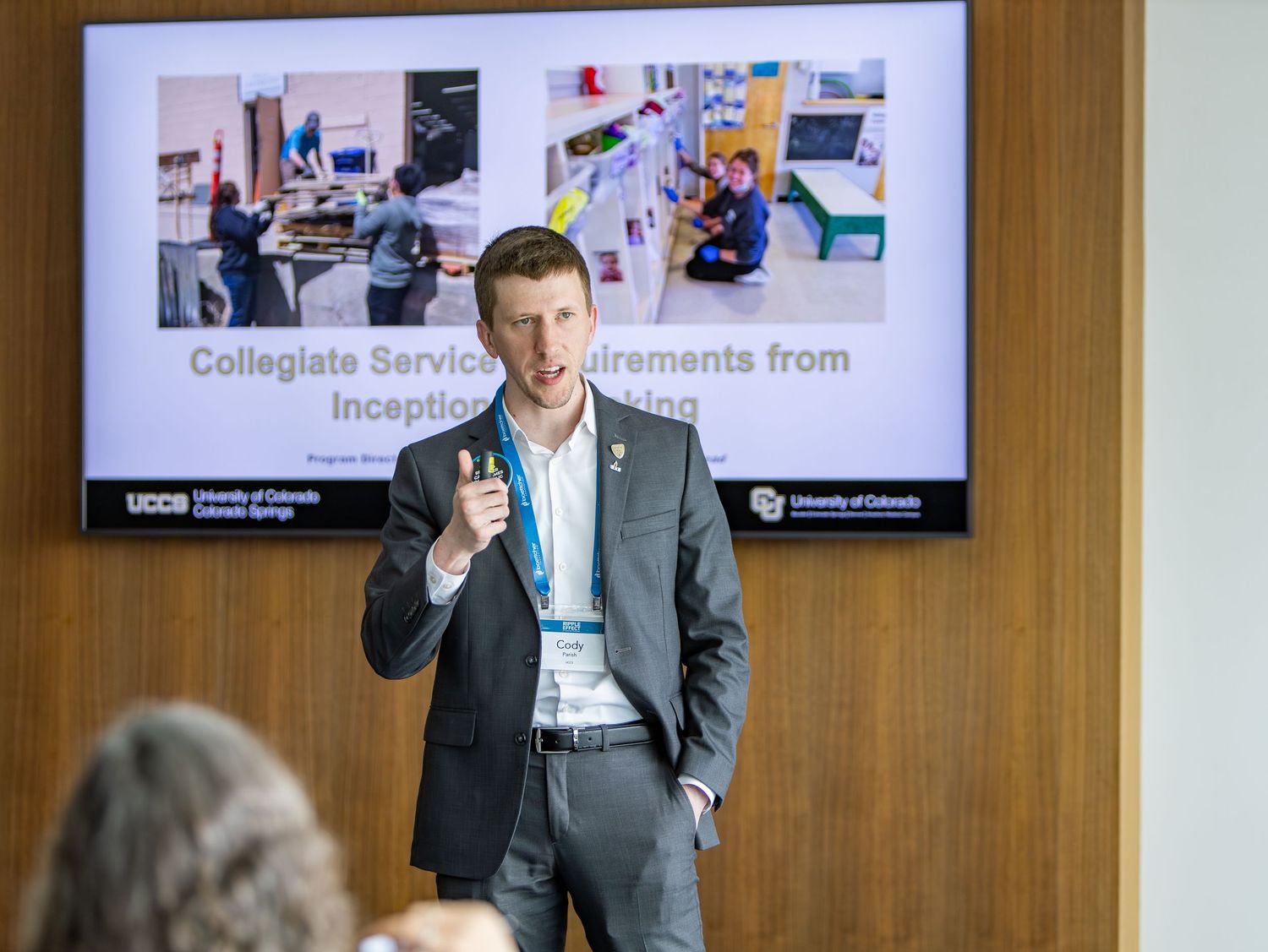 Business professional in a gray suit gestures while speaking during a corporate presentation.