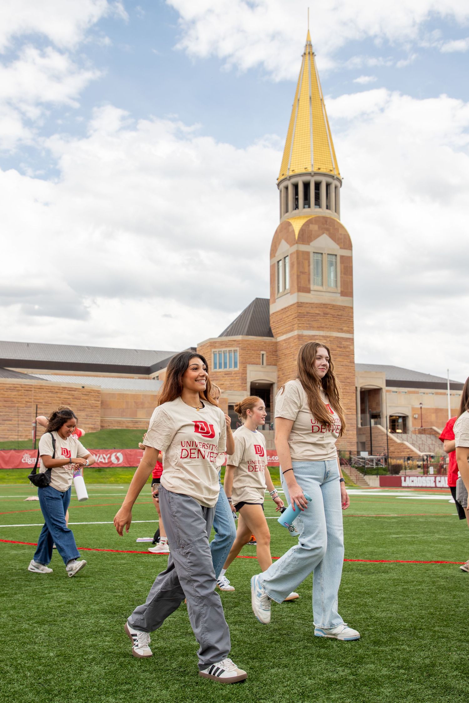 Students walk on athletic field during University of Denver orientation with iconic gold-spired building in background.