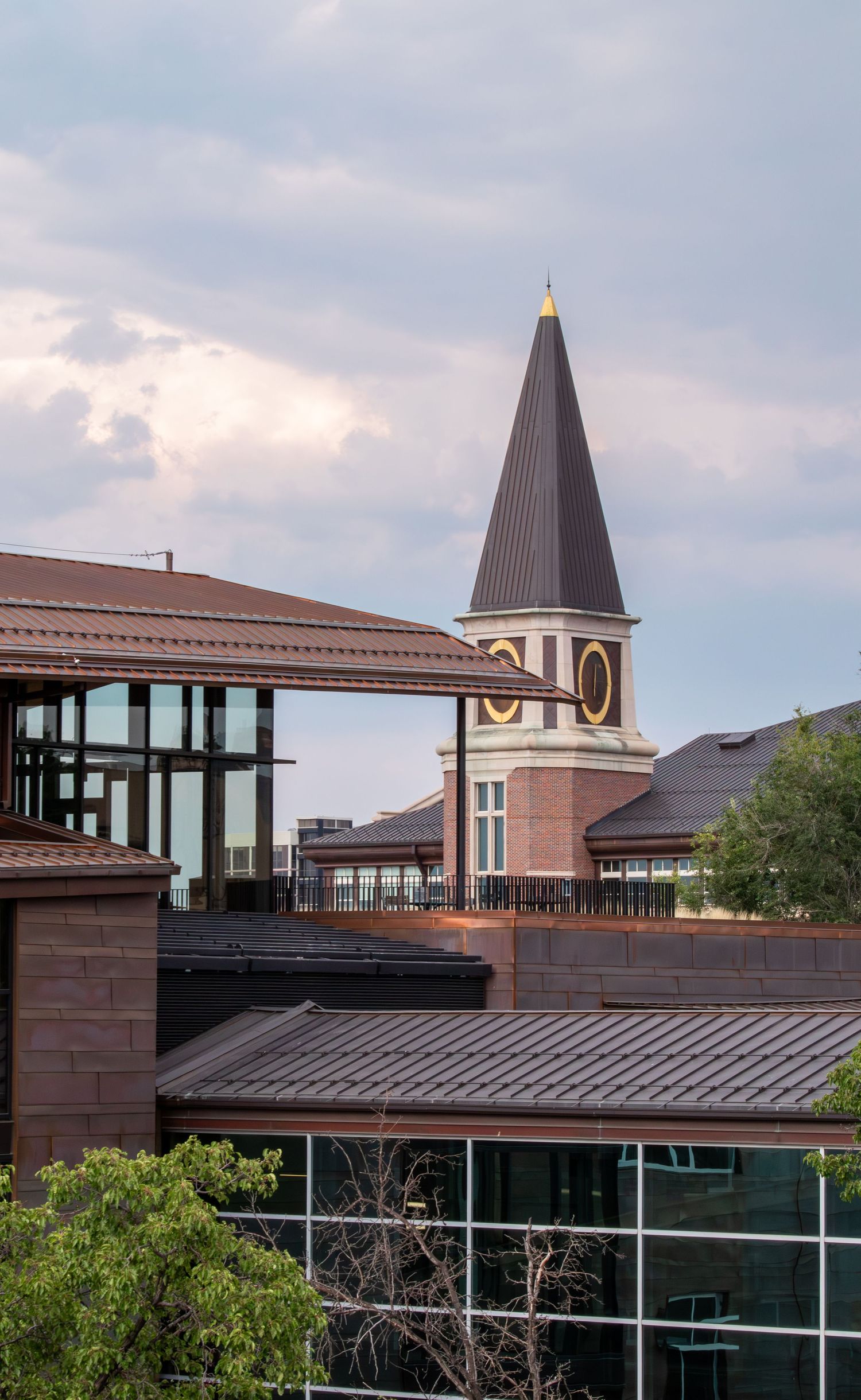 Historic brick clock tower with dark spire rises above modern buildings under a cloudy sky.