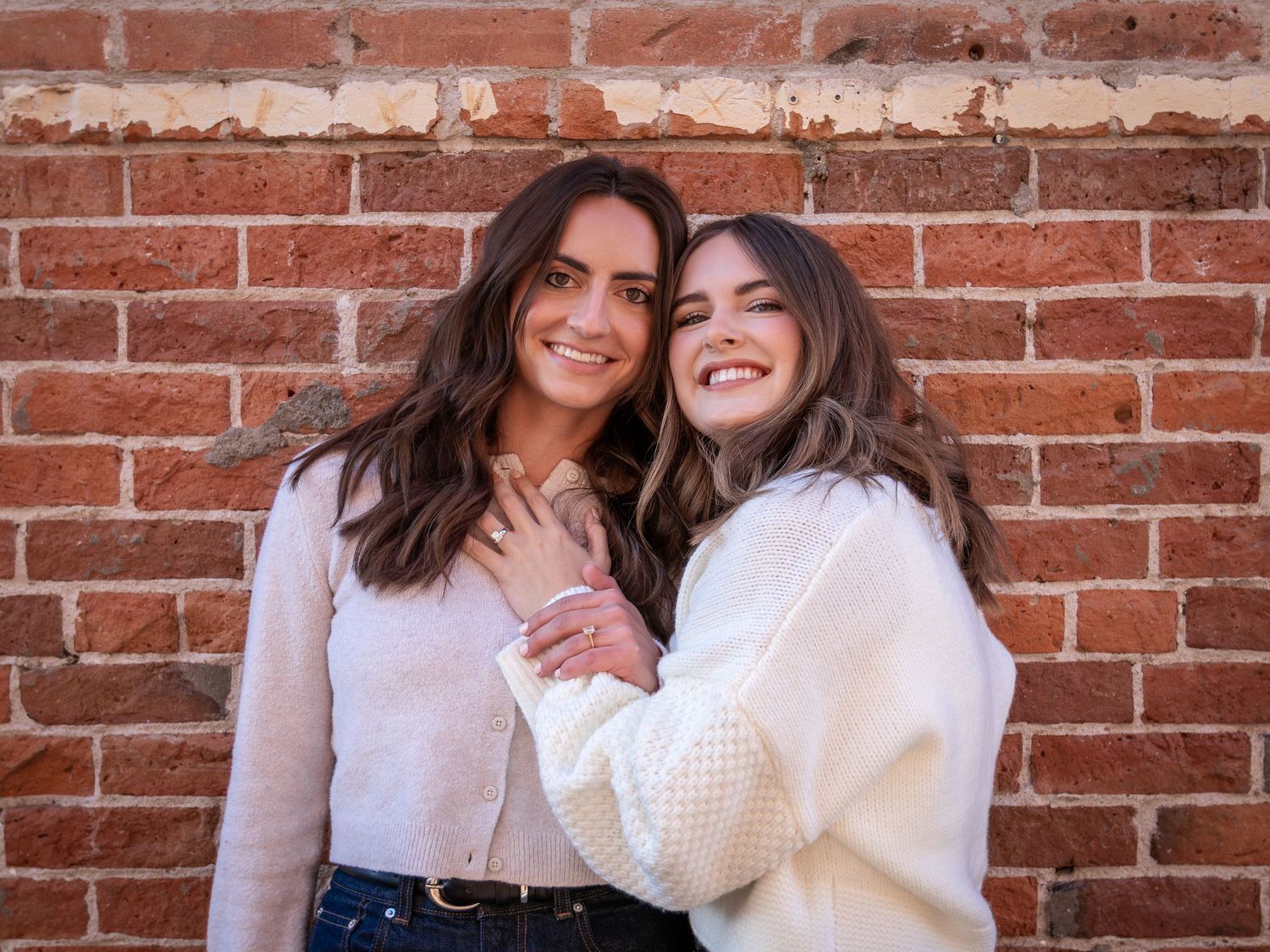 Two friends in white sweaters smile and embrace while posing against a rustic brick wall background.