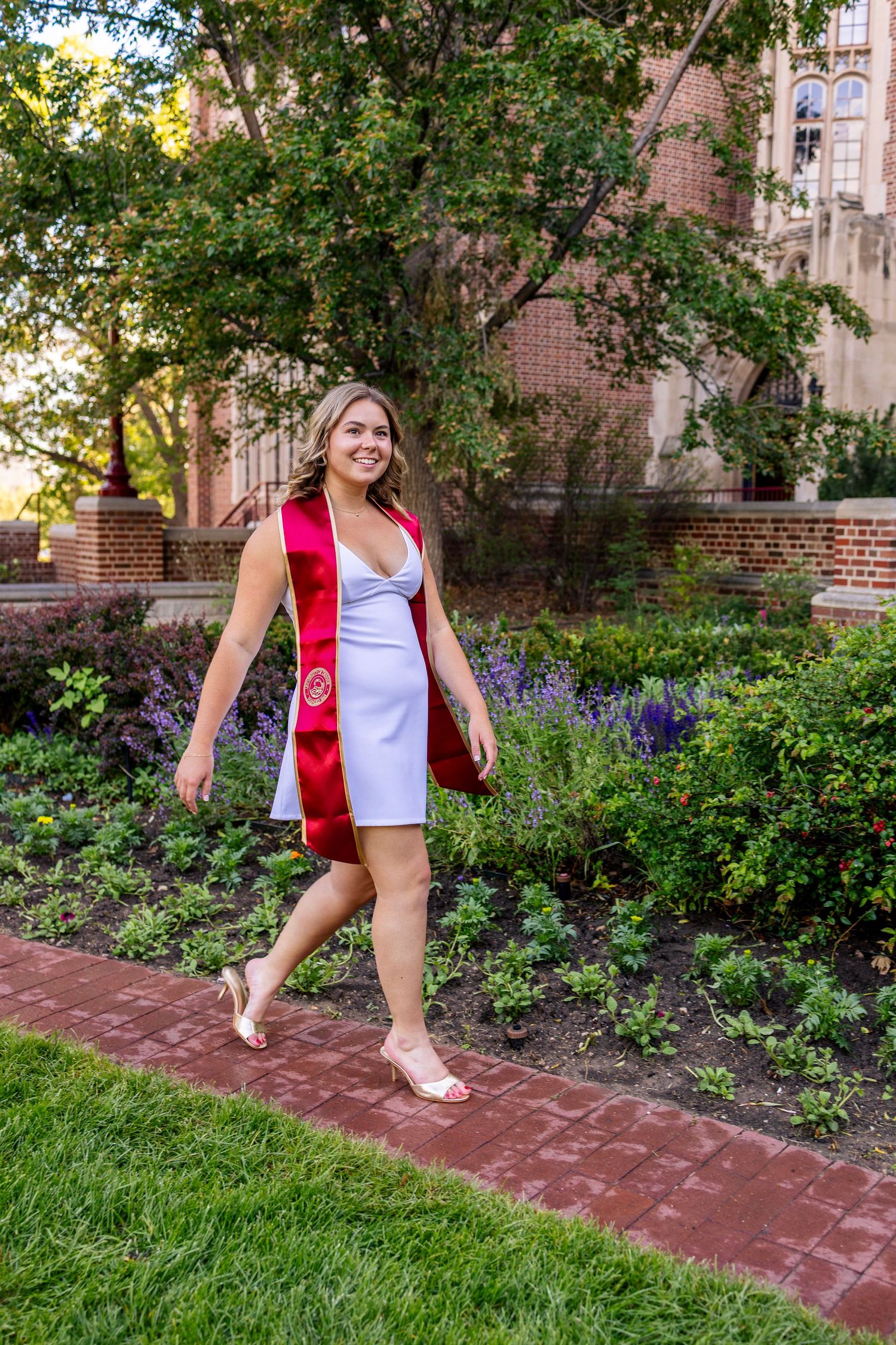 Smiling female graduate in white dress and red sash walks along brick path beside colorful garden near brick building.