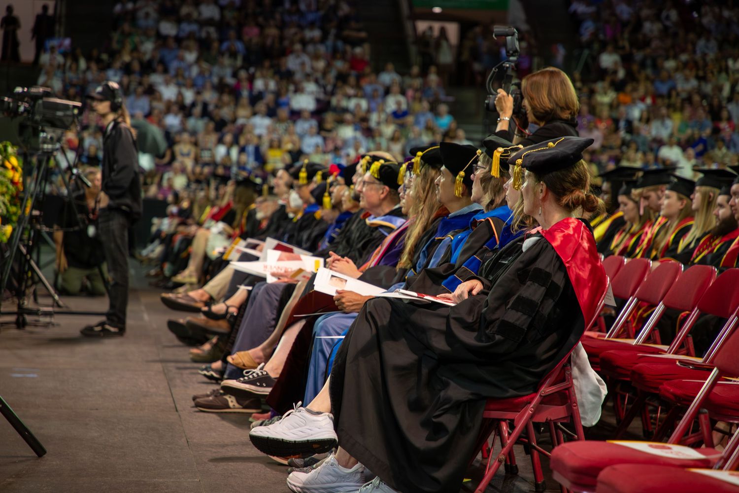 Graduates in caps and gowns sit in rows of red chairs during an indoor commencement ceremony.