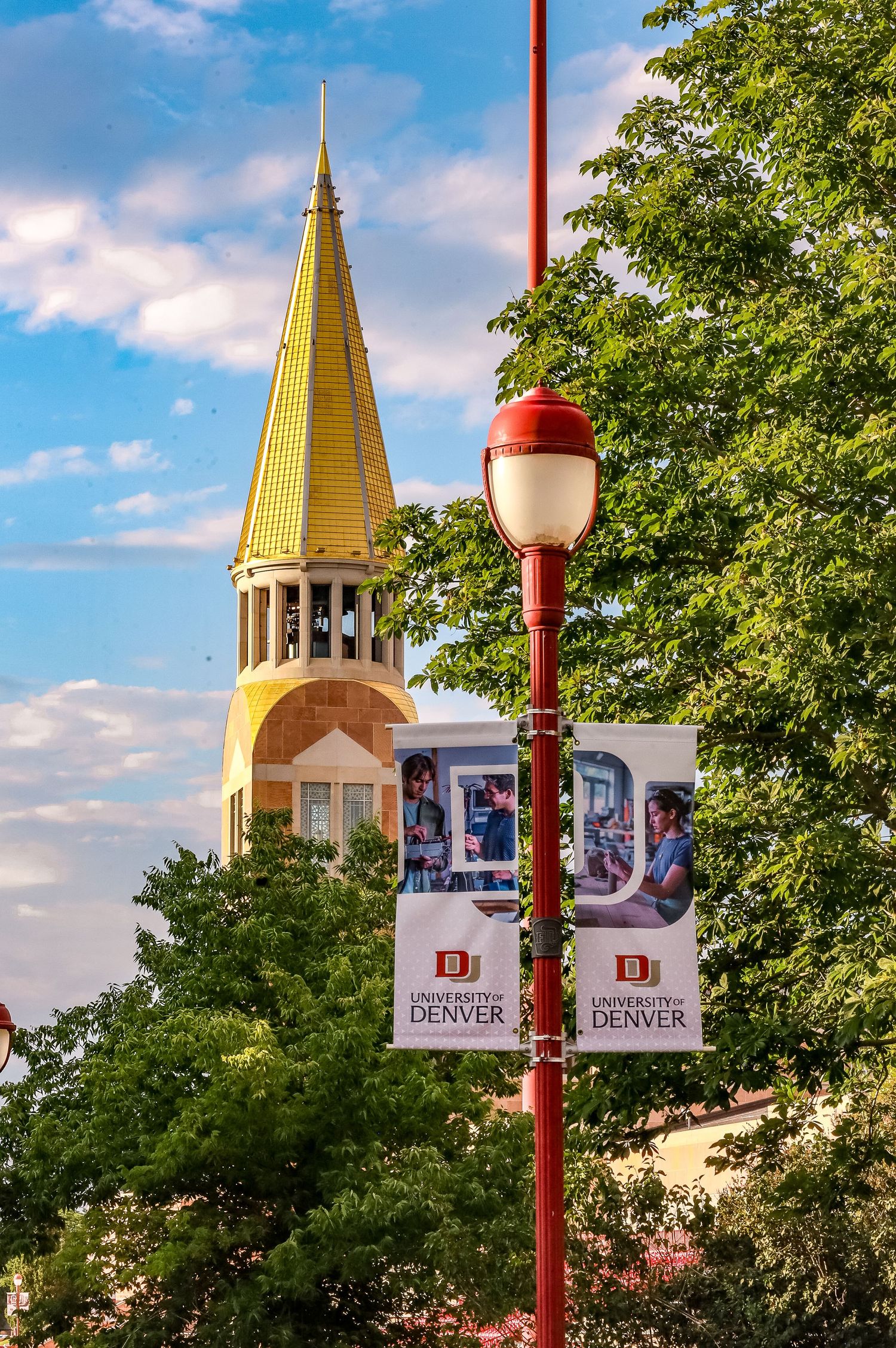 Golden-domed clock tower of the University of Denver campus rises above trees against a blue summer sky.