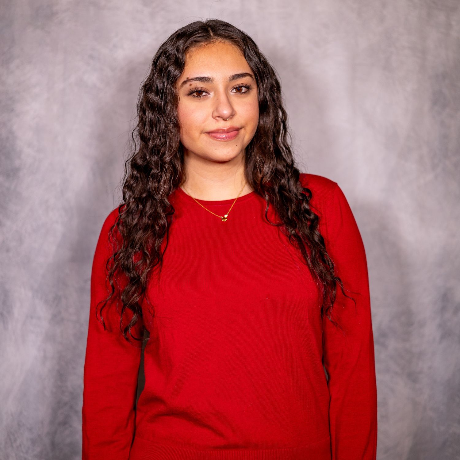Young woman with long curly dark hair wearing a red long-sleeve shirt and gold necklace poses for a portrait.