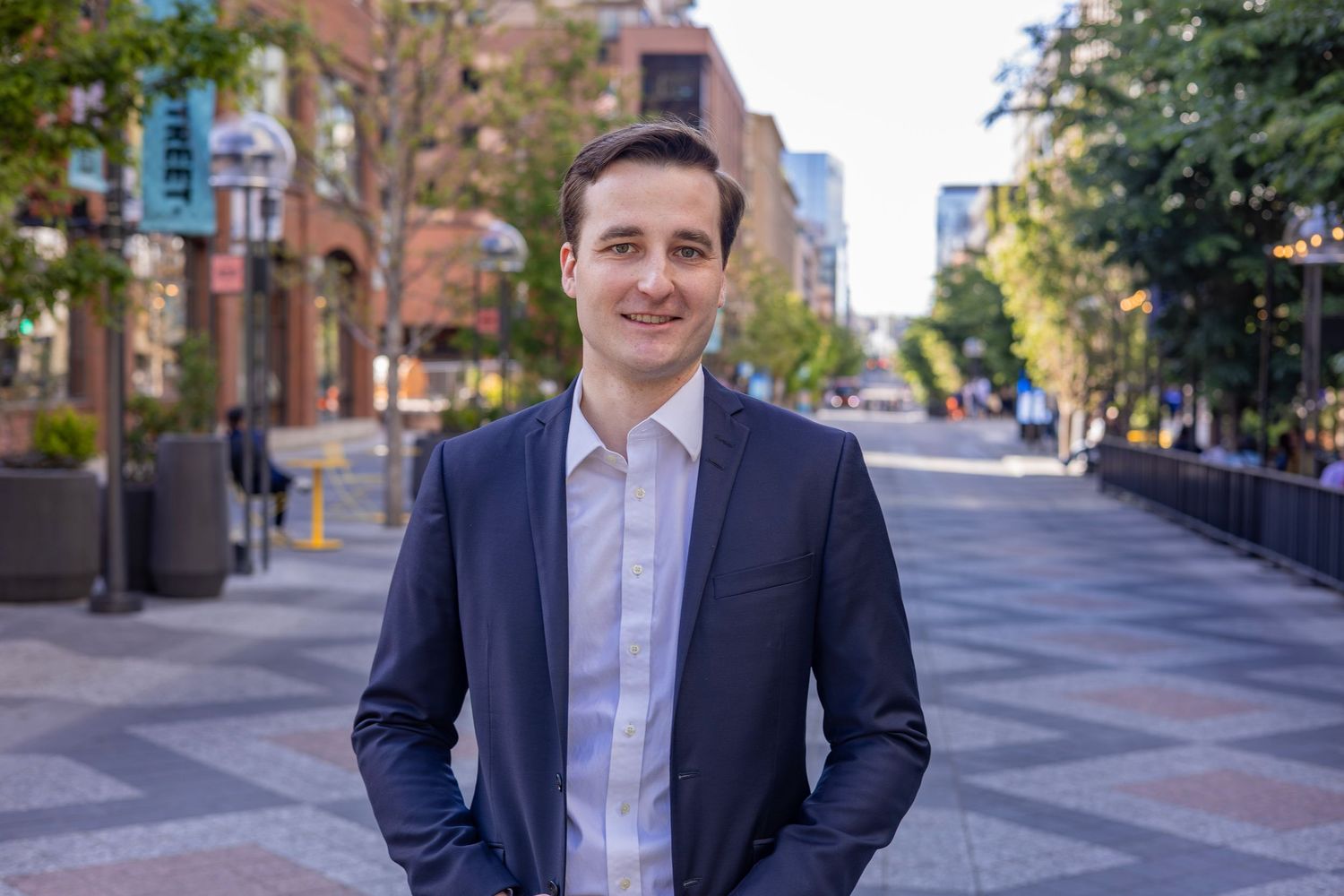 Young professional man in navy suit smiling on urban brick pedestrian street with trees and city buildings.