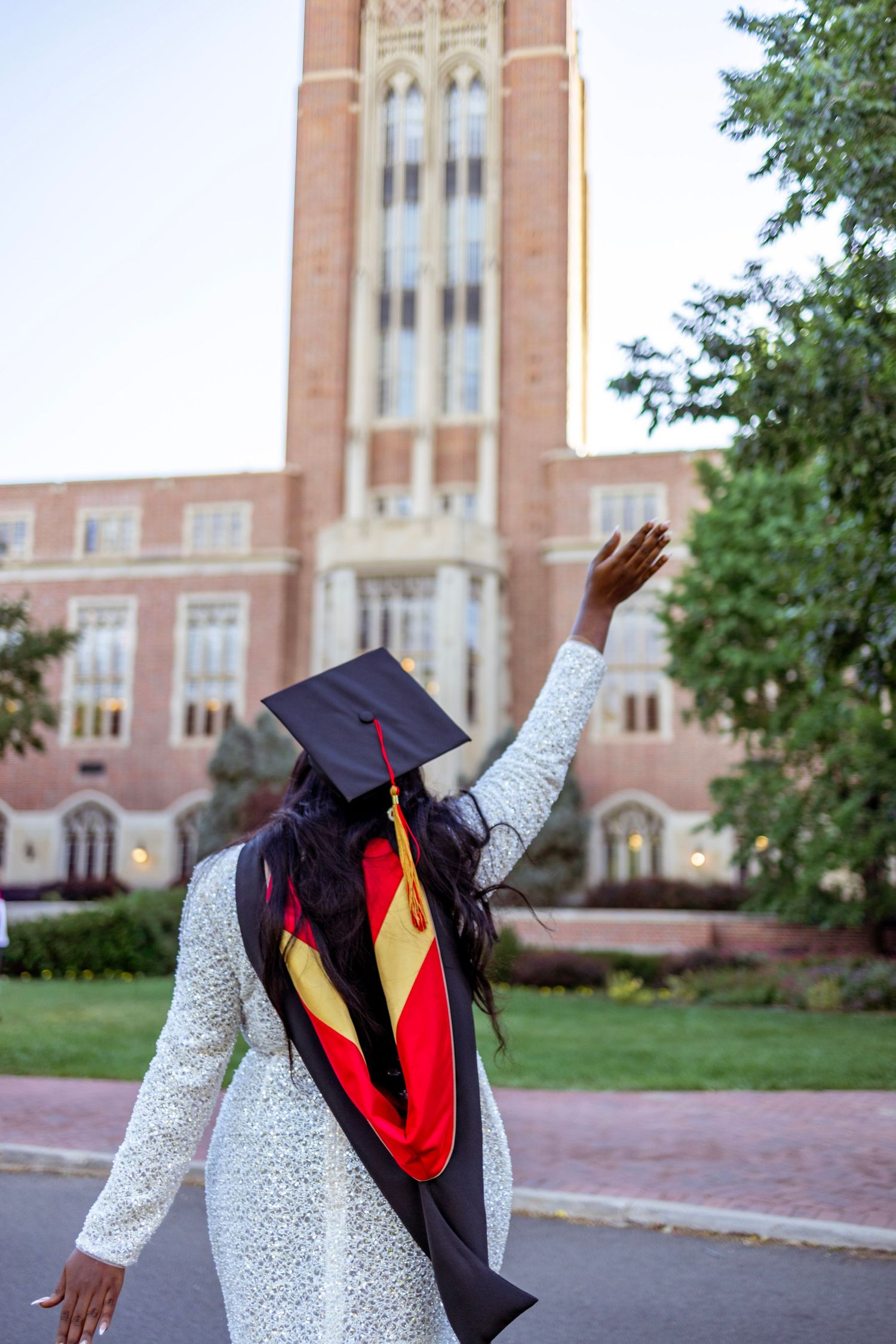 Graduate in cap and gown raises hand triumphantly in front of a brick university tower building.