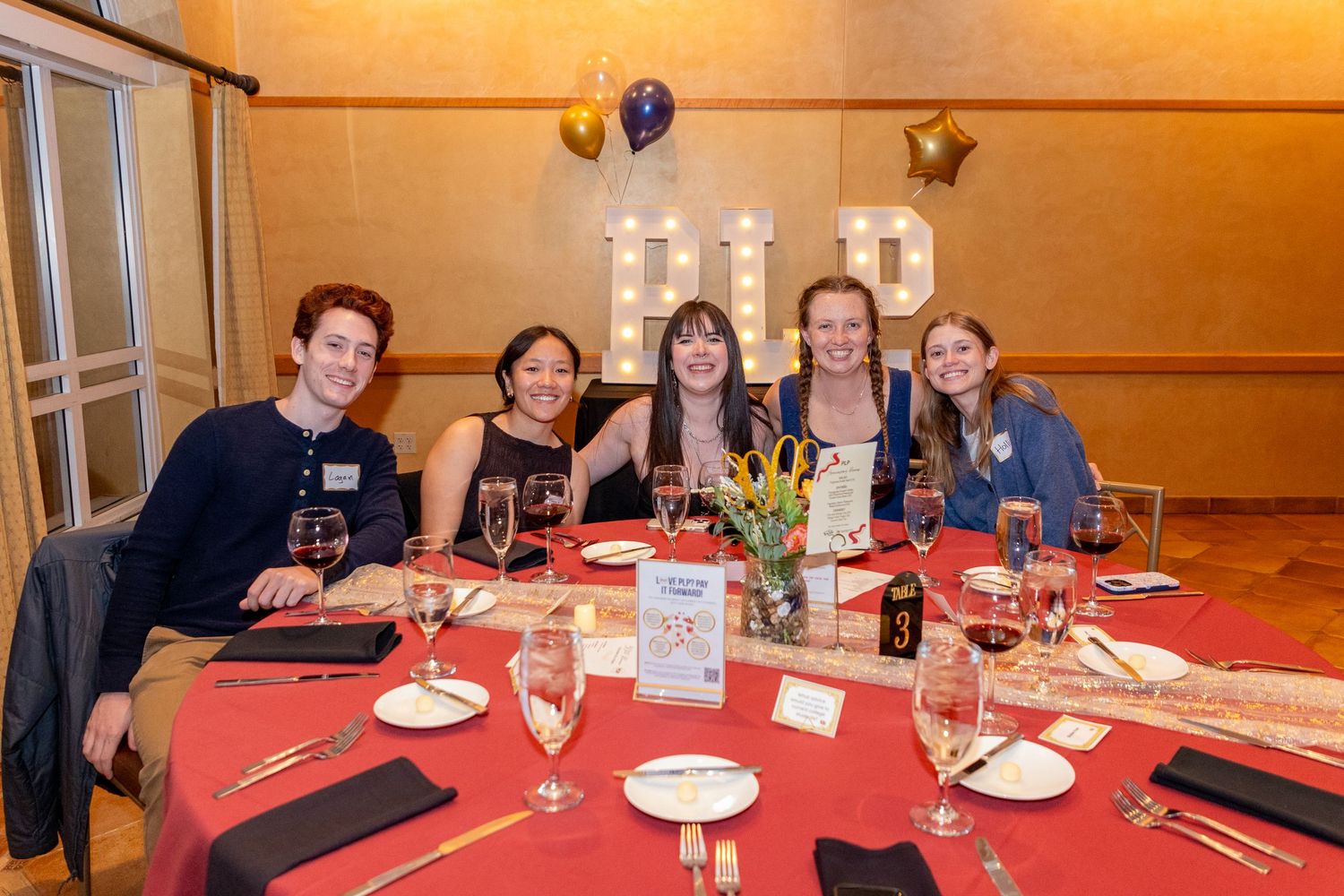 A group of people celebrate at a formal dinner event with red tablecloths, wine glasses and festive decorations.