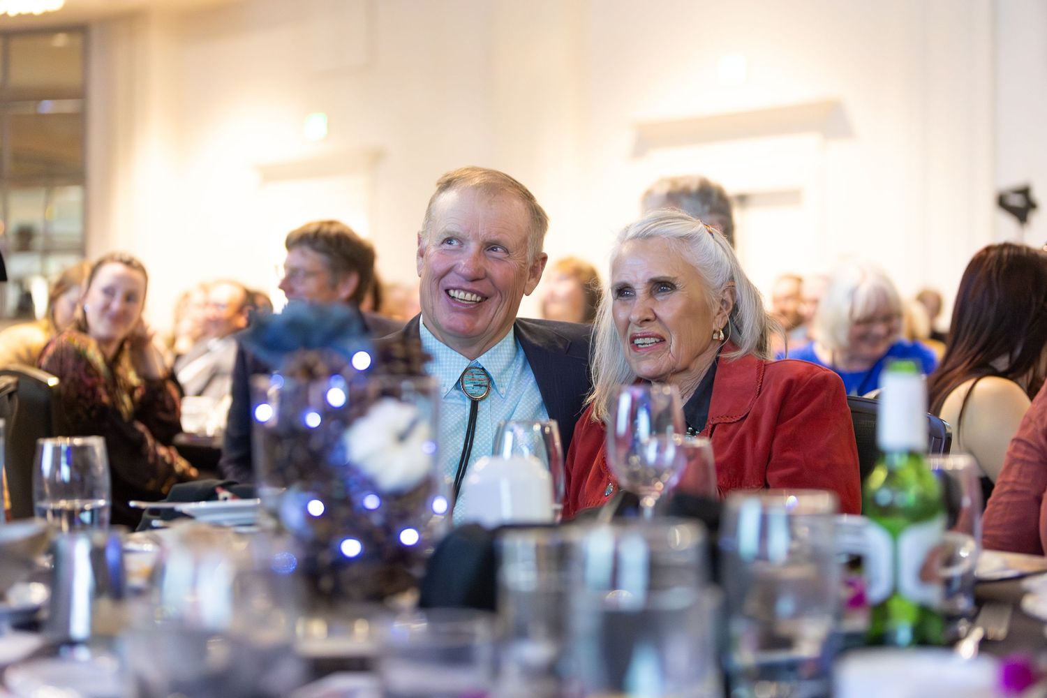 People enjoying a lively social gathering at a dining table with drinks and laughter in a bright indoor setting.