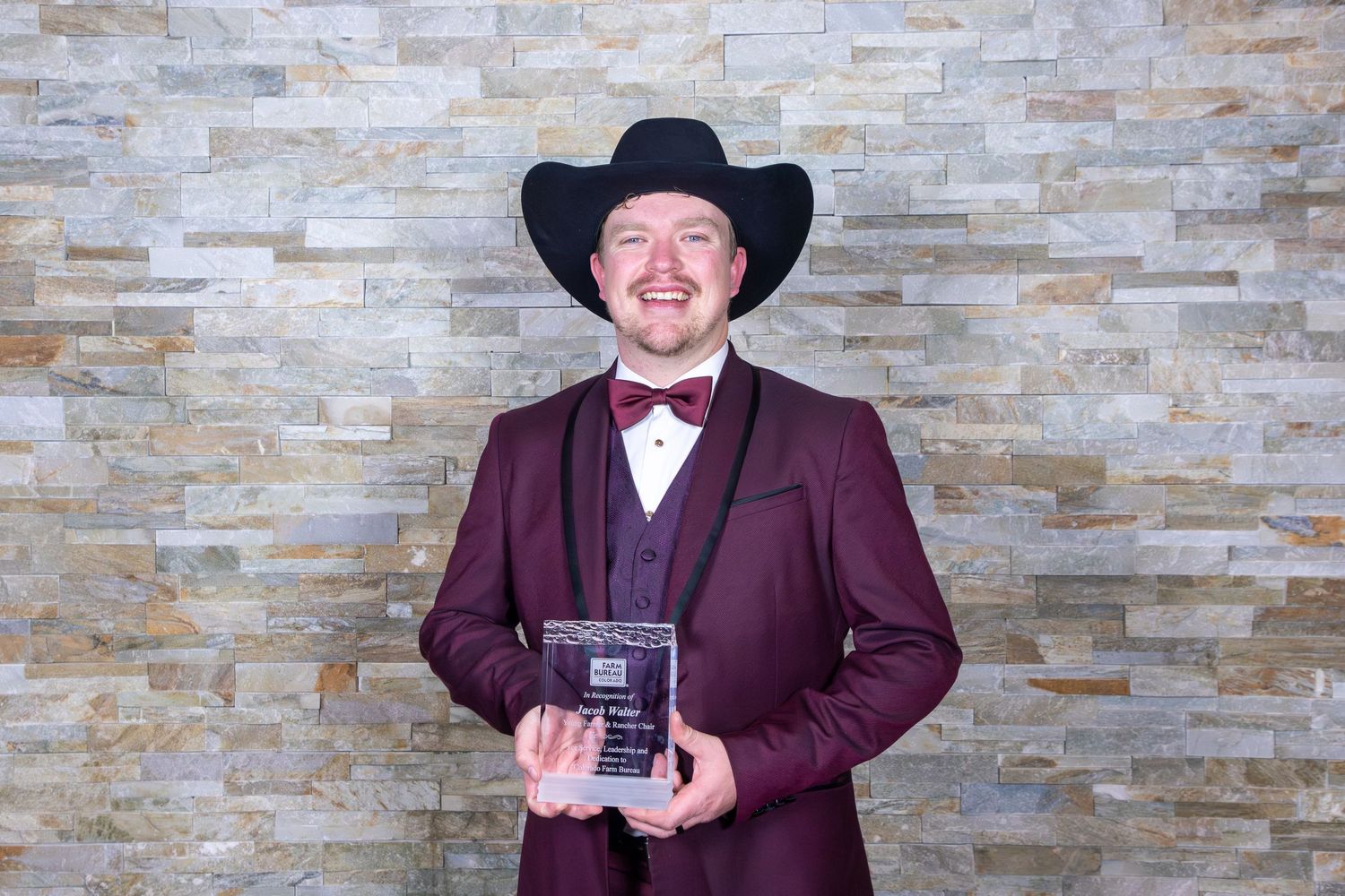 A person in a burgundy suit and black cowboy hat poses with an award against a stone wall backdrop.
