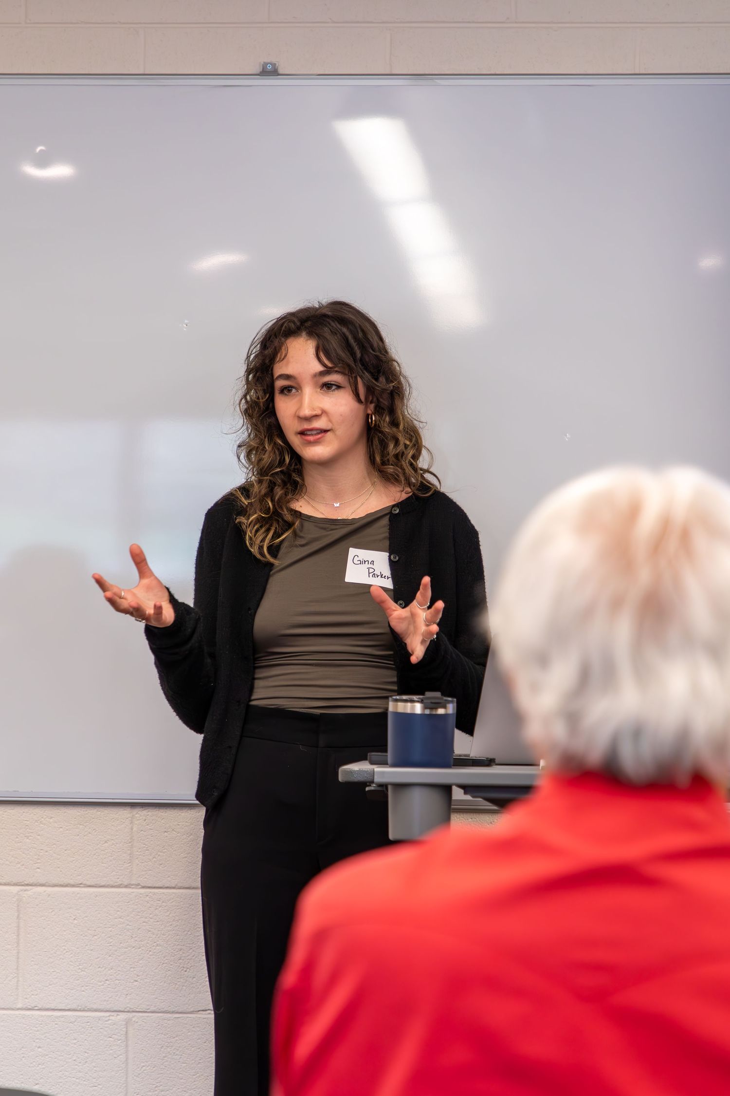 Speaker gesturing while giving a presentation in front of a whiteboard.