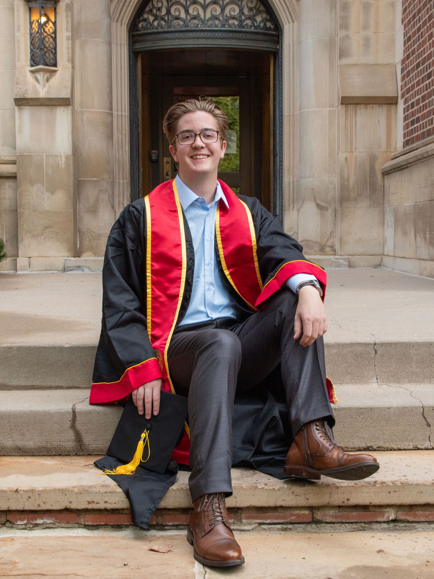 Graduate in academic regalia sits on stone steps of historic building entrance.