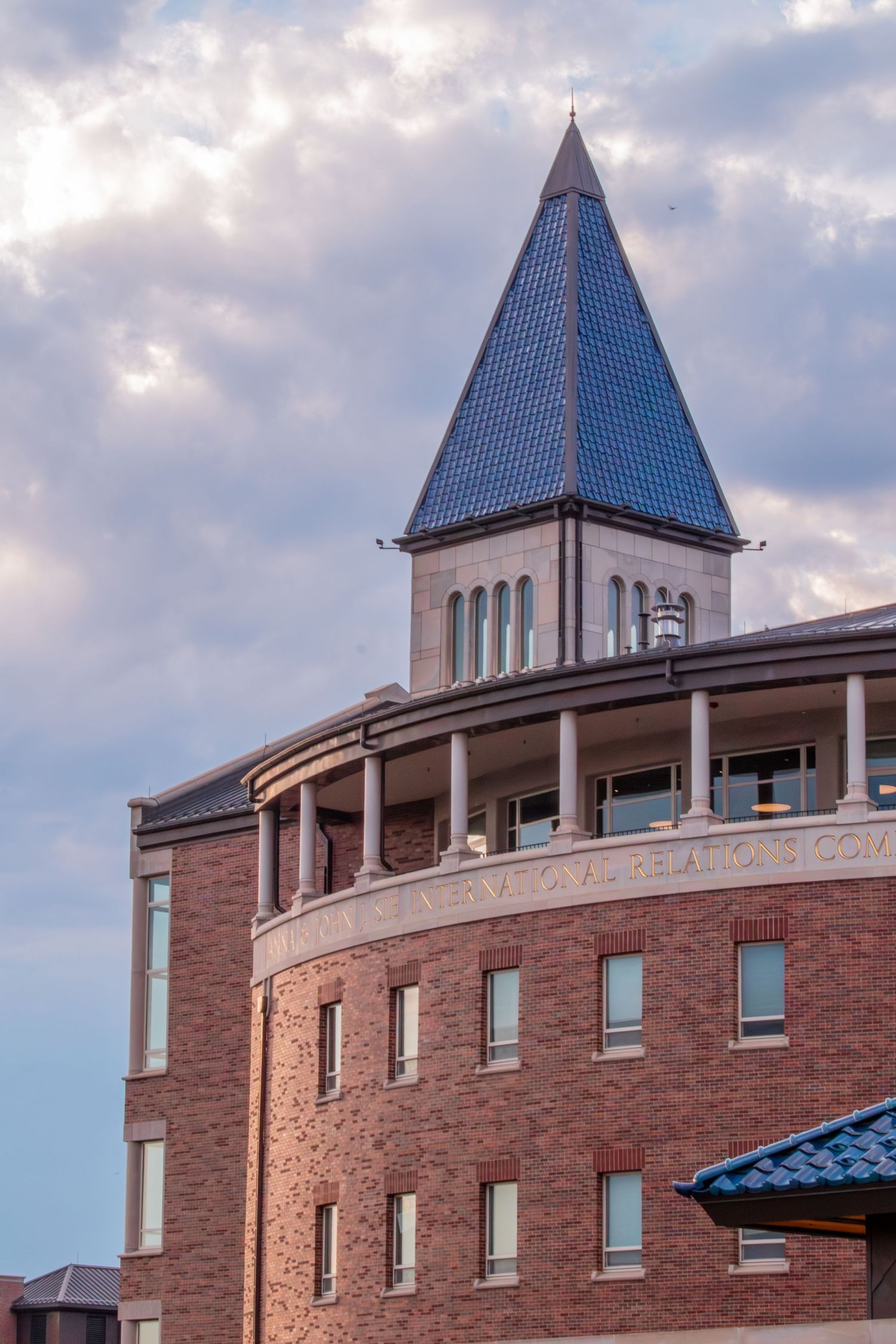 Historic brick building with blue pyramid spire tower against cloudy sky, featuring arched colonnade and large windows.