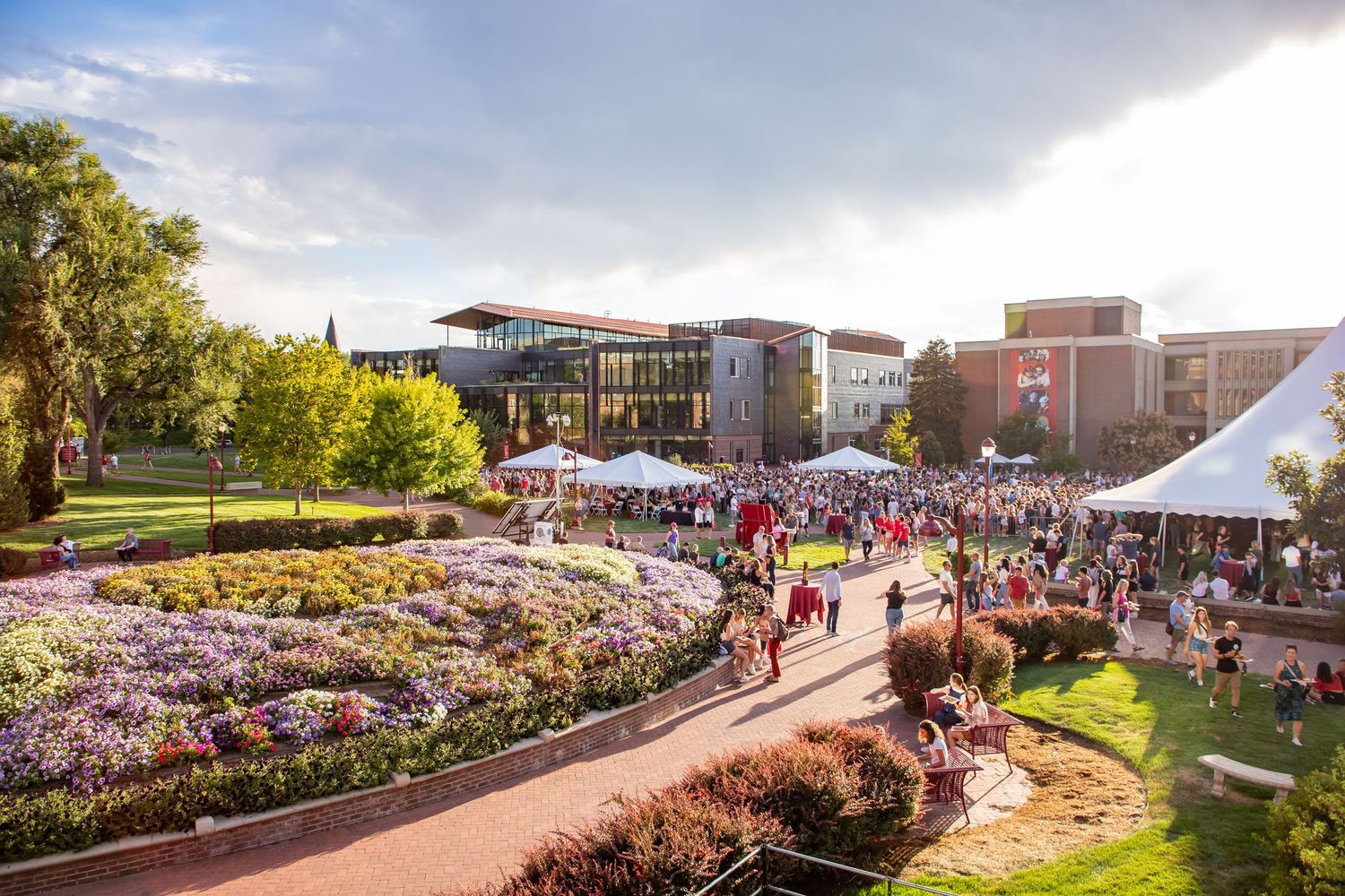 Aerial view of a vibrant university campus event with crowds, white tents, colorful gardens, and modern buildings at sunset.