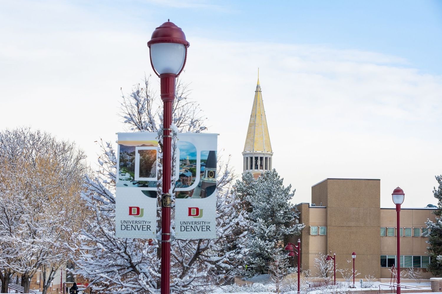 A scenic winter campus view with snow-covered trees, red lampposts, and academic buildings under a cloudy sky.