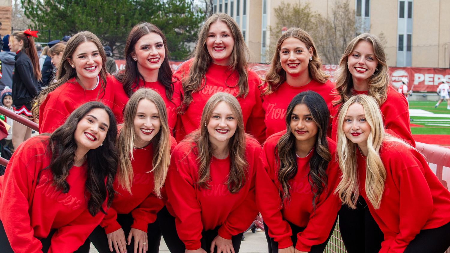 A group of students wearing matching bright red sweaters pose together outdoors on a college campus.