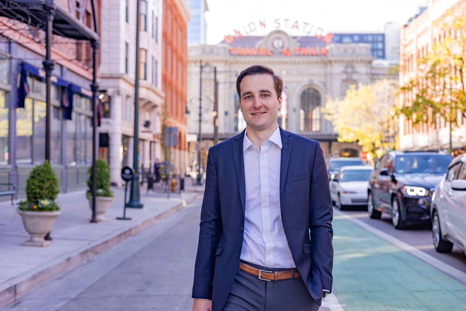 Professional man in navy suit smiling on urban street with green bike lane and historic building backdrop.