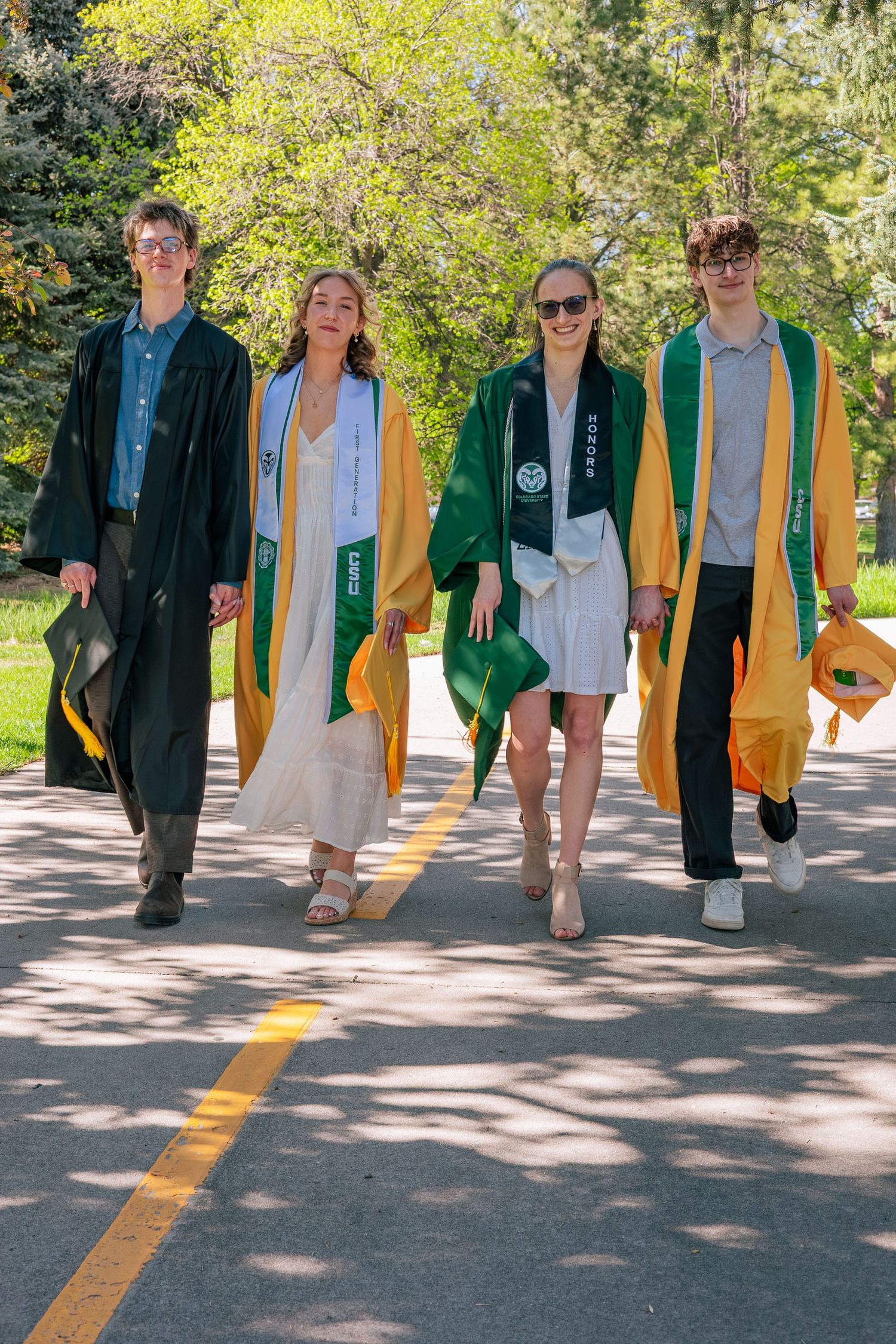 Four college graduates in green and gold graduation robes walk together outdoors on a sunny day.