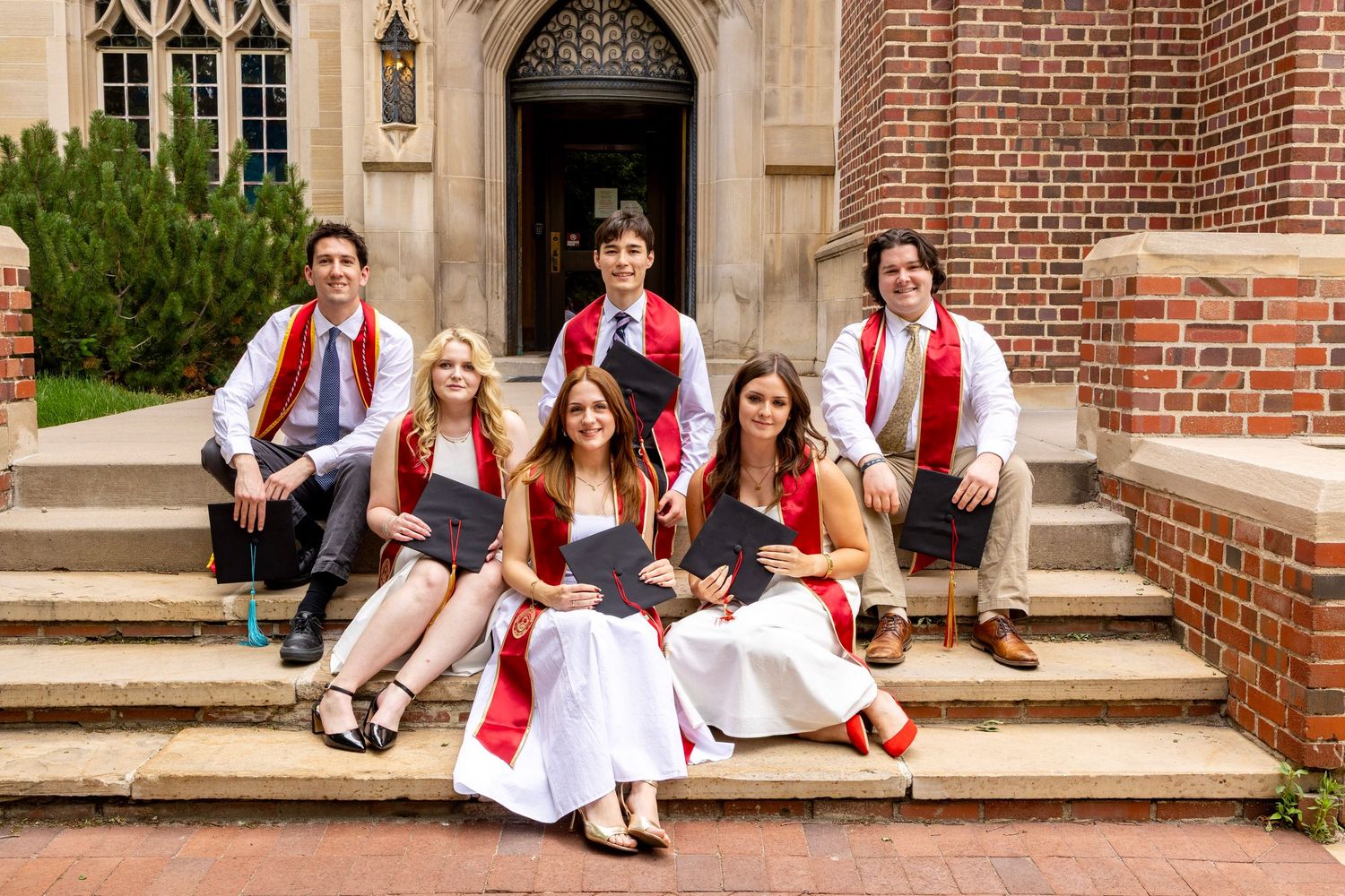 Seven graduates in red and white regalia pose on brick steps outside a stone building, smiling and holding diplomas.