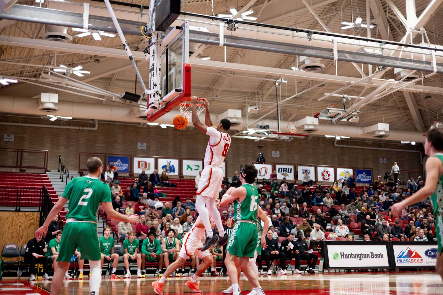 Basketball player dunks over defenders in a packed red and white college gymnasium during an intense game.