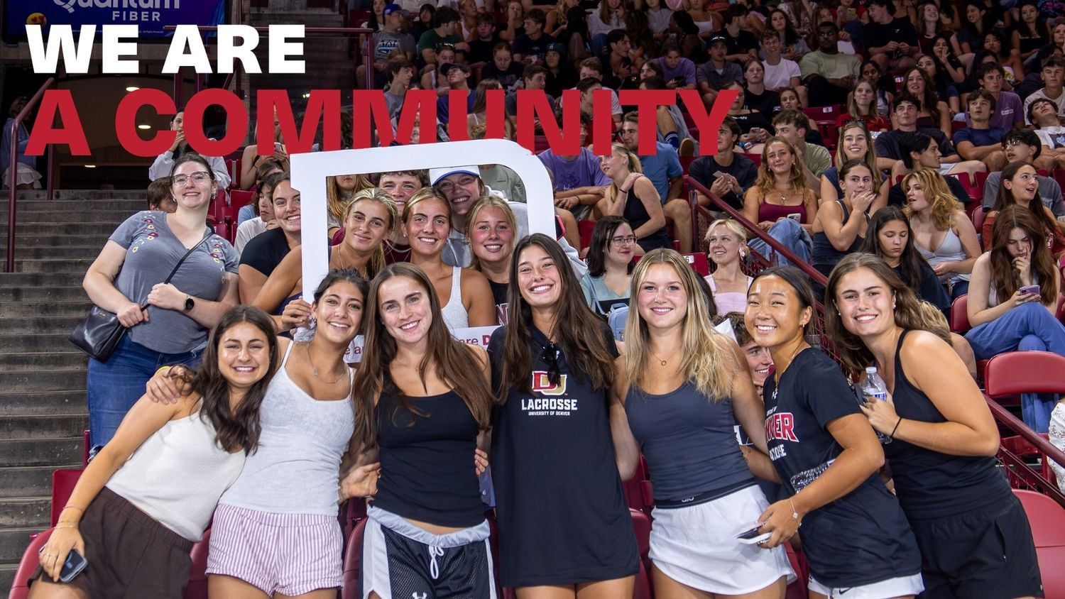 College students gather for a community photo at a sports arena, smiling together under a We Are A Community banner.