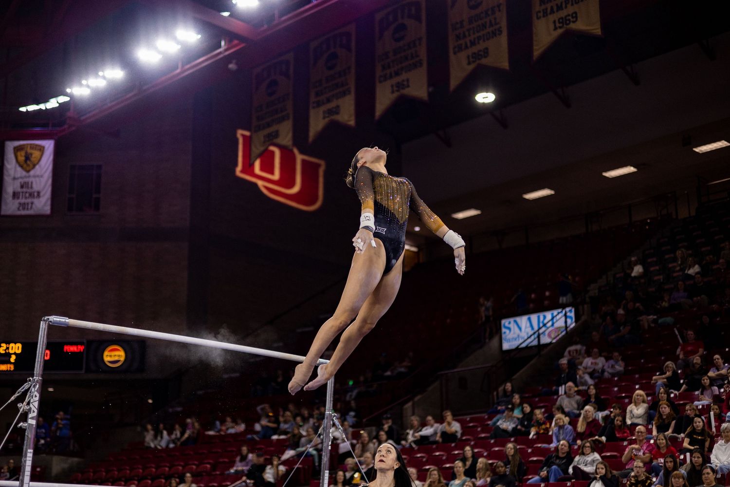 Female gymnast performs a release move on uneven bars in a packed university arena with championship banners.