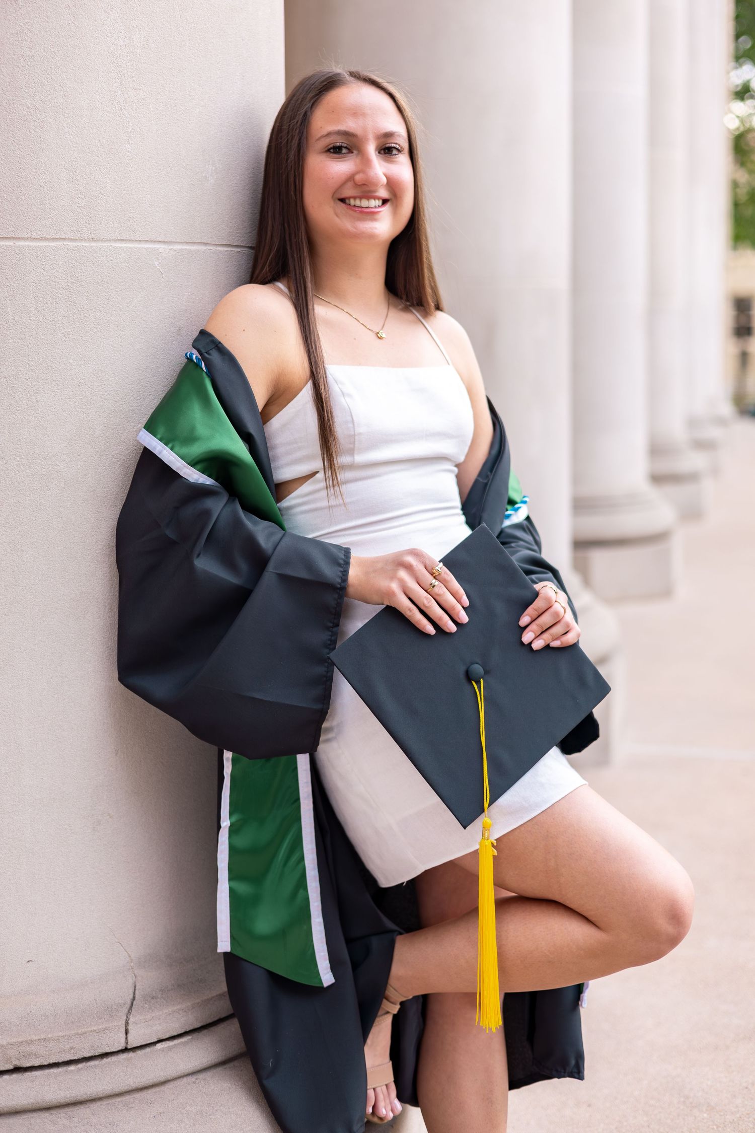 Smiling graduate leaning against column in green and black graduation gown, holding cap with yellow tassel.