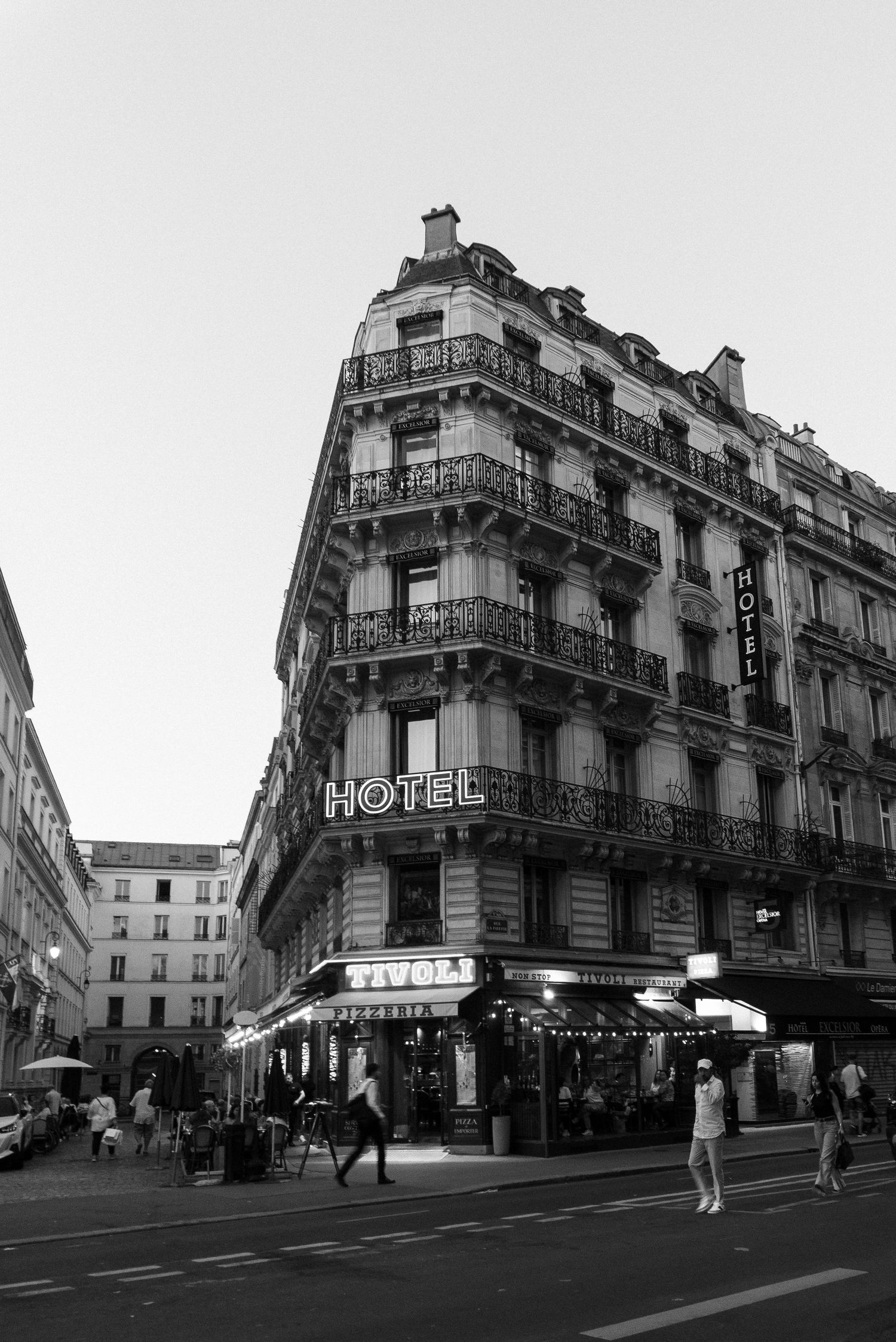 Black and white photo of a classic Parisian hotel building with ornate balconies and a corner cafe on a busy street.