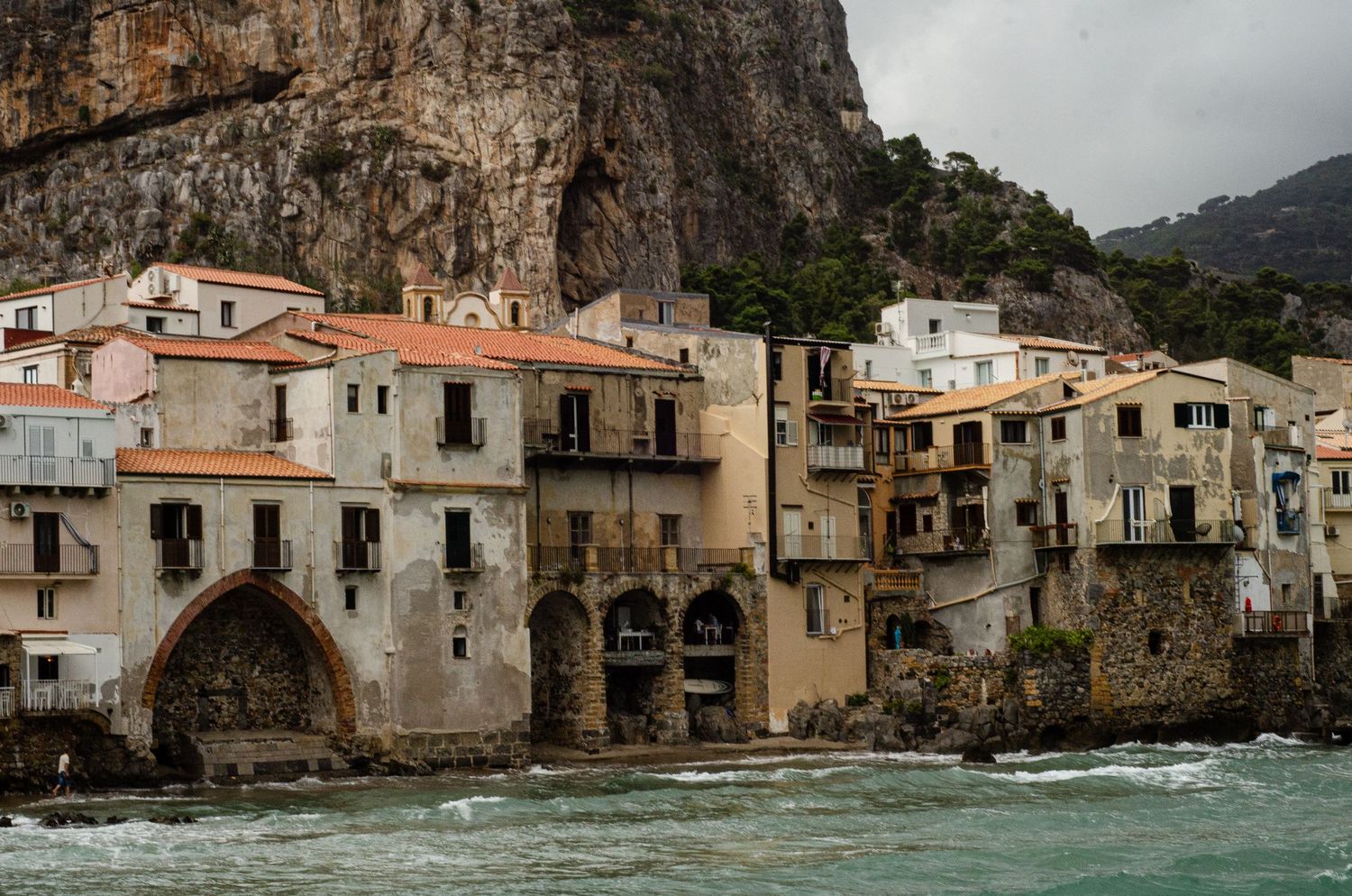 Historic seaside houses with white walls and terracotta roofs built into cliffs along the Mediterranean coast in Cefalu, Sicily.